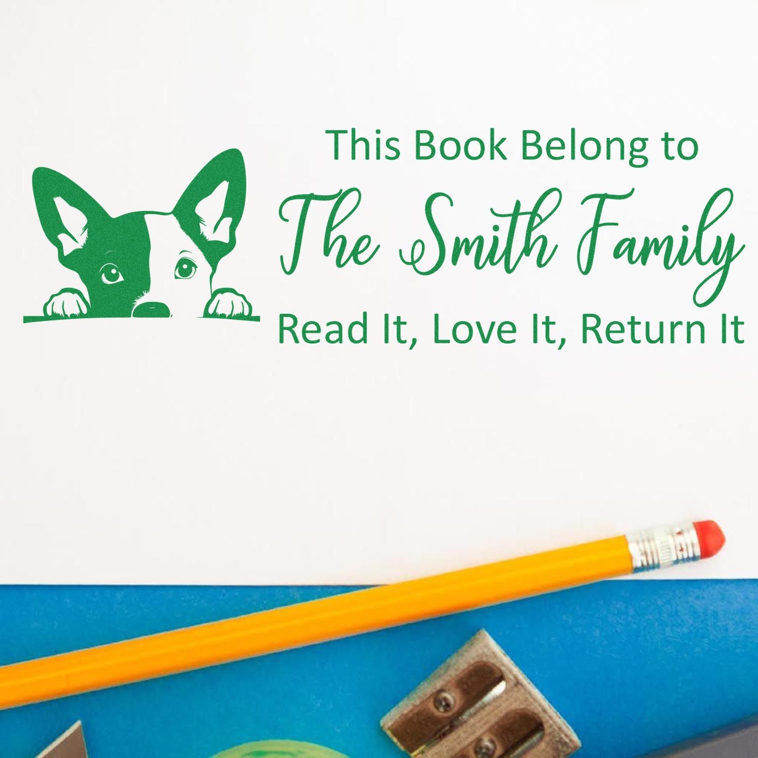 Green custom library stamp with dog and This Book Belongs to The Smith Family on white, plus yellow pencil and blue background.