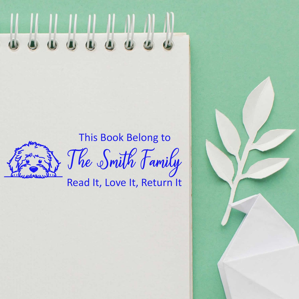 Blue personalized book stamp on spiral notebook, The Smith Family with dog icon, on mint green background and white paper leaf.
