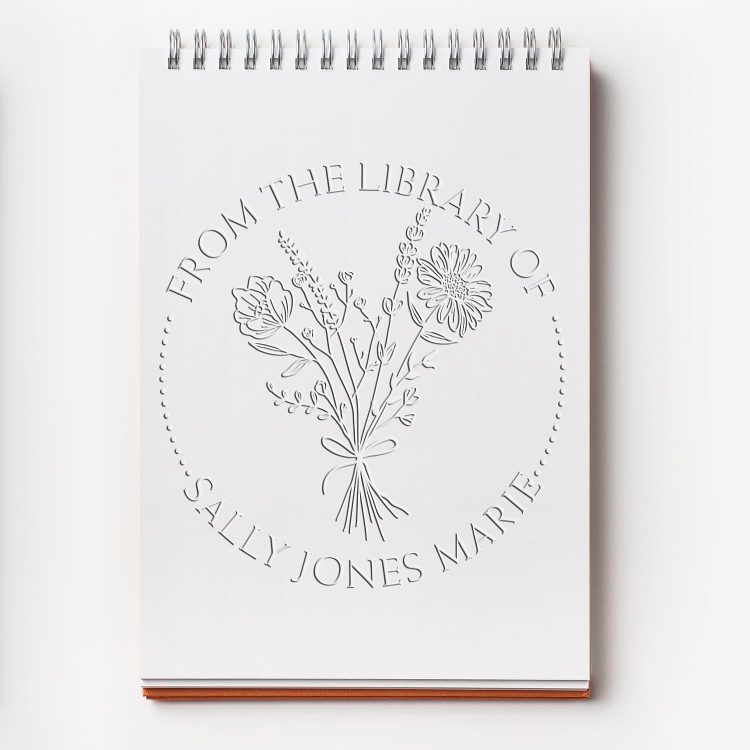 White spiral-bound notebook with an embossed floral library stamp seal in gray, reading “From the Library of Sally Jones Marie.”