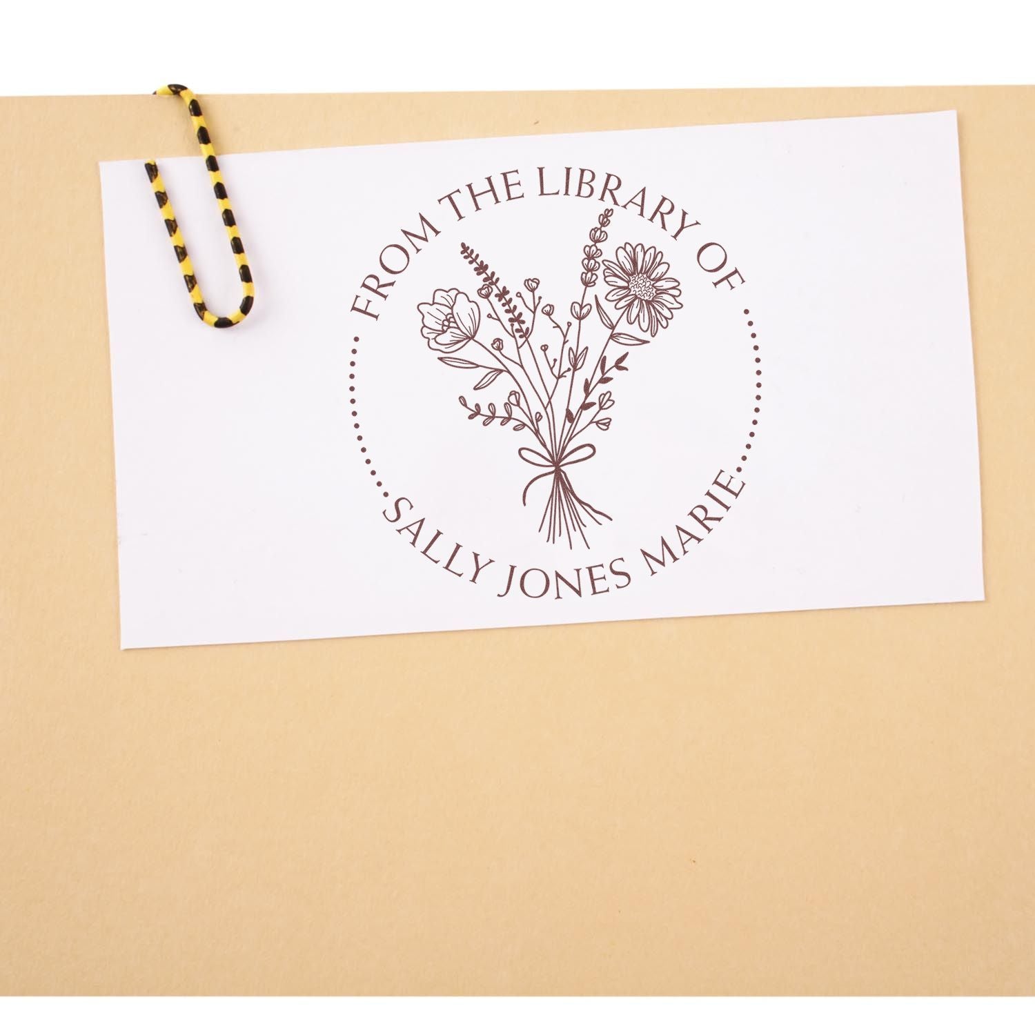 Custom brown floral library stamp and seal on white card, clipped with a yellow-and-black paperclip against beige background.