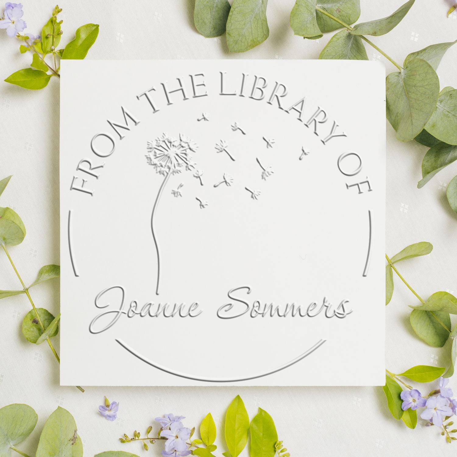 White embossed stamp and seal with dandelion design and From the Library of Joanne Sommers on a green leafy floral background.