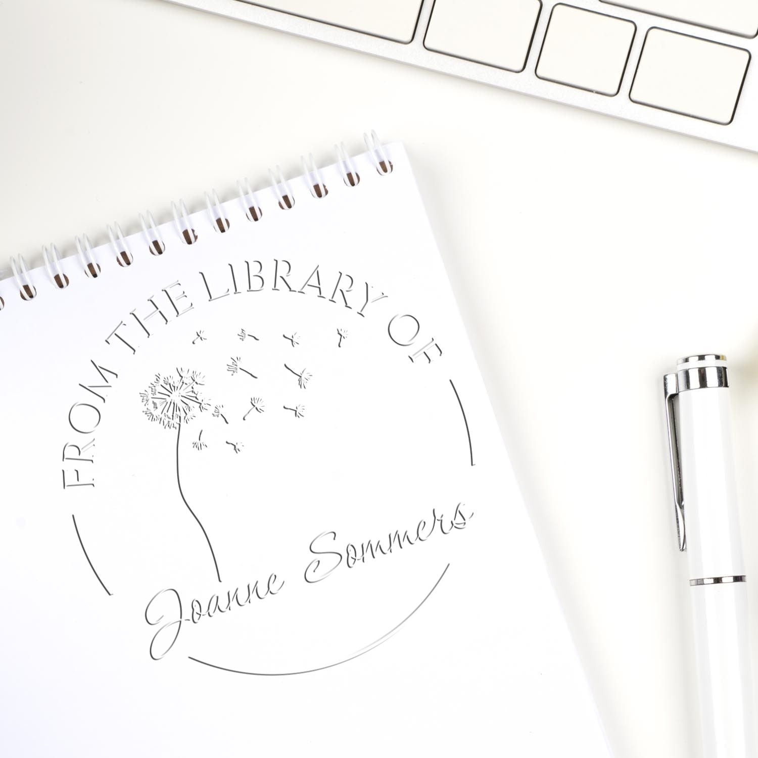 White custom library stamp and seal with dandelion design, embossed on notebook, beside a silver pen and keyboard.
