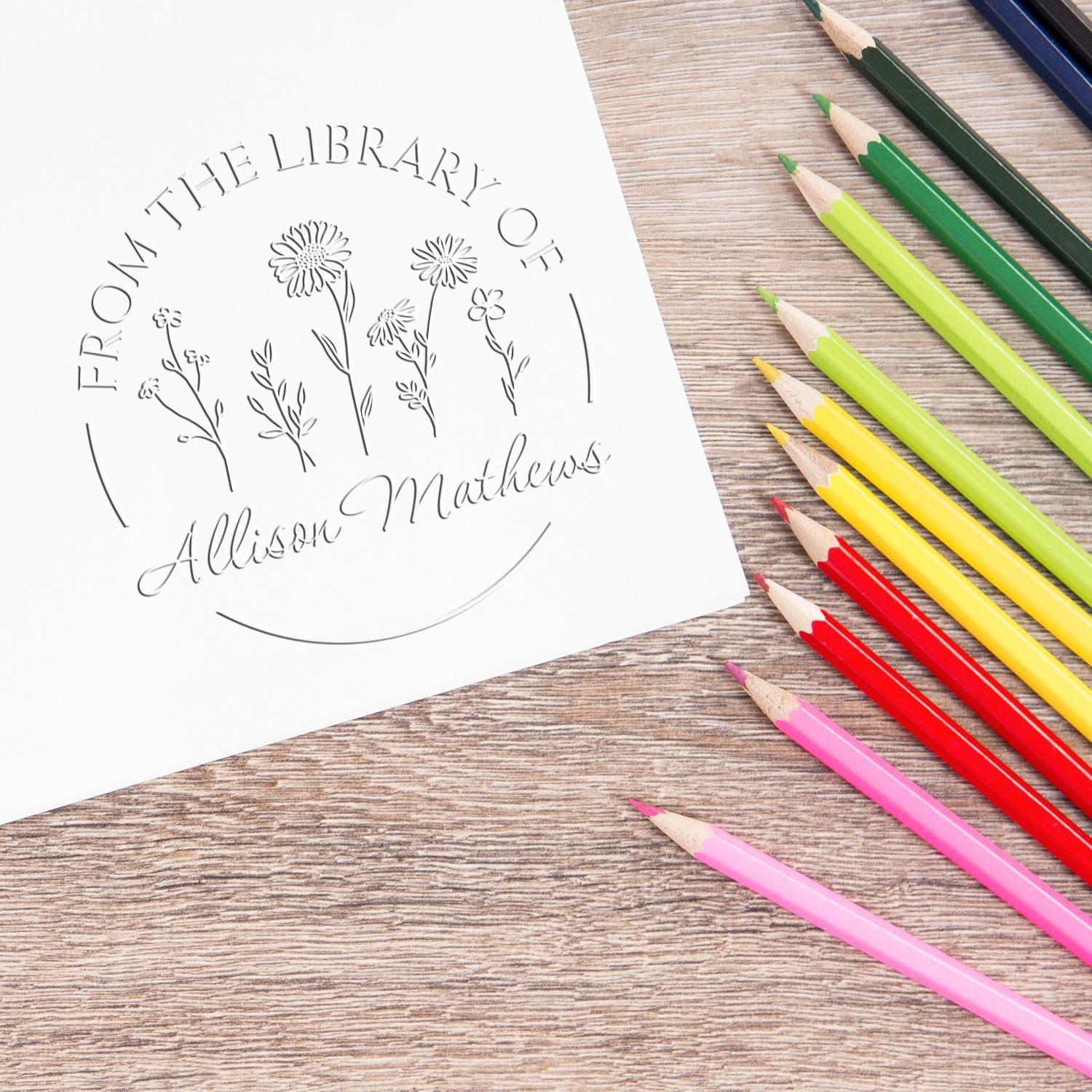 Custom library stamp and seal embossed on white paper with floral design, beside colorful pencils on a wood desk.