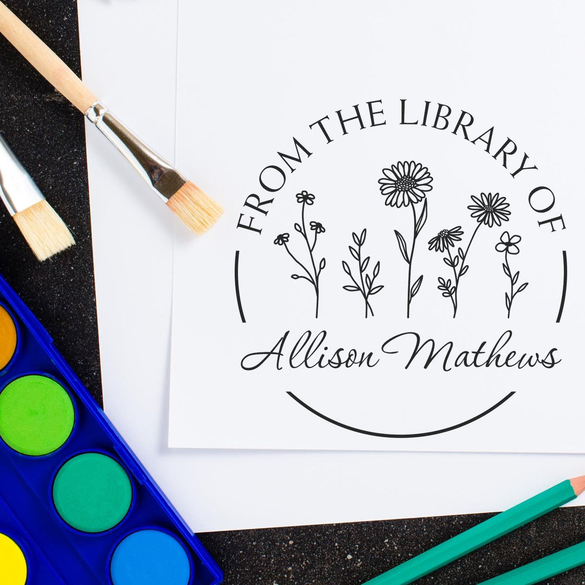 Personalized library stamp seal with black floral design and Allison Mathews name on white paper, beside bright paintbrushes and paints