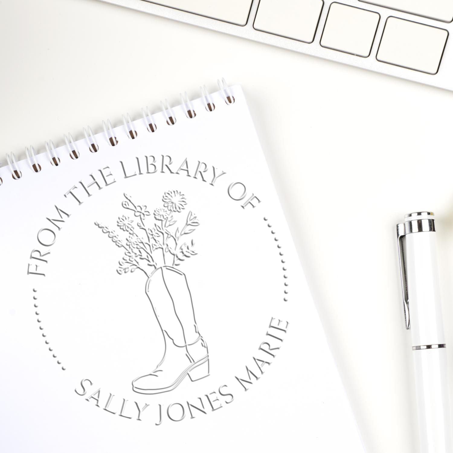 White custom library bookplate stamp and seal design with floral cowboy boot, beside a silver pen and keyboard.