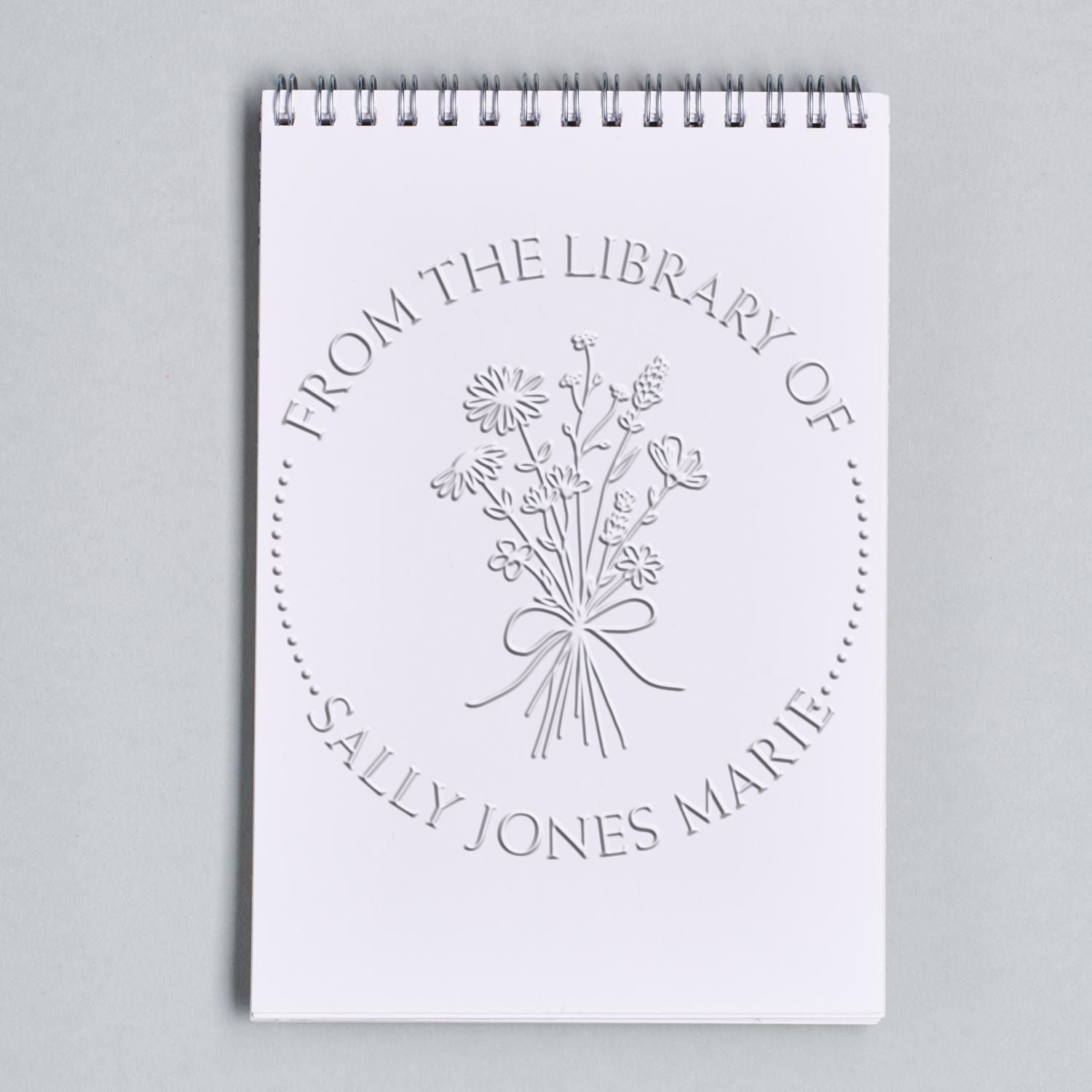 White spiral notebook with an embossed floral library stamp seal, custom name text, and silver spiral binding on gray.
