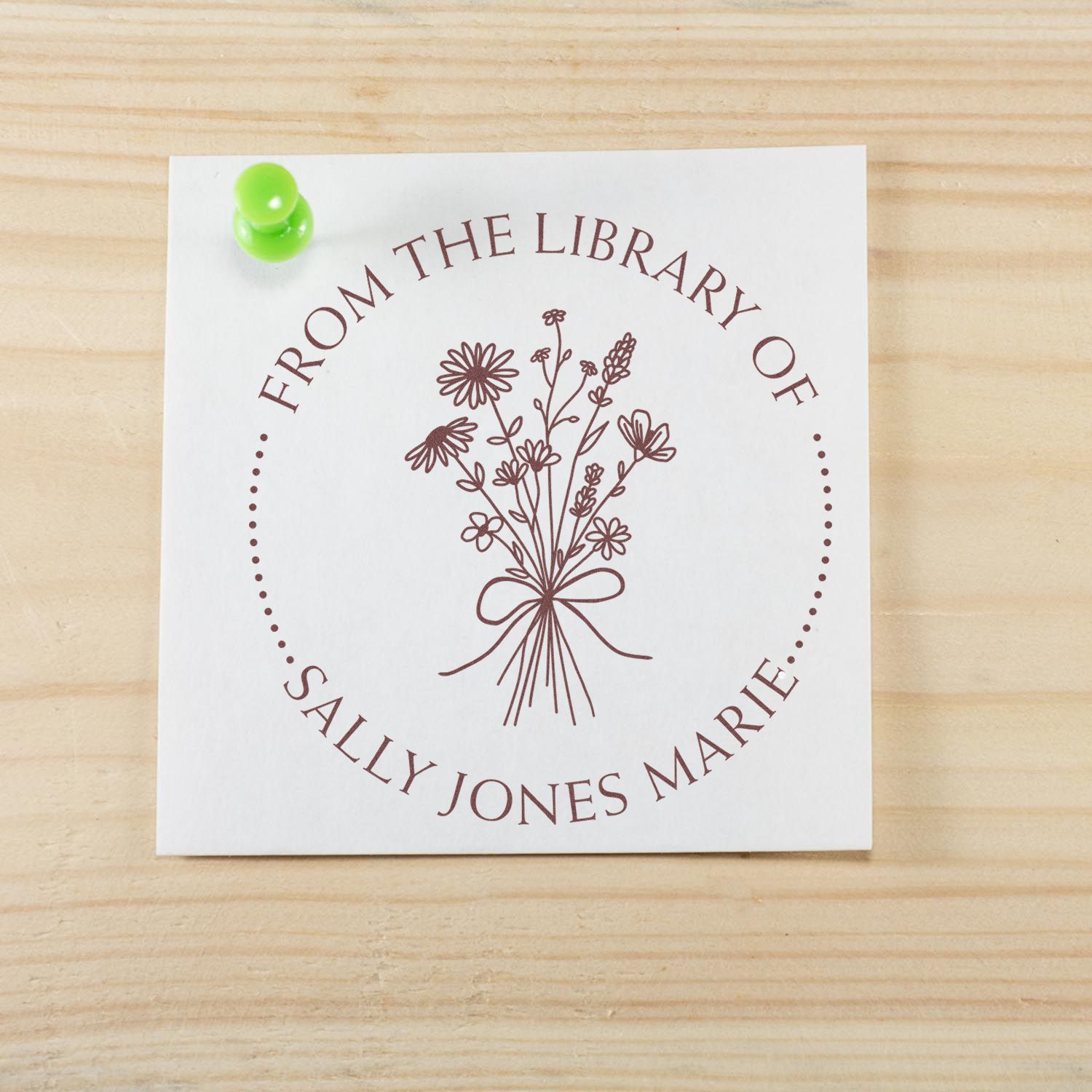 Custom brown return address stamp seal with floral bouquet, pinned to a white card by a green tack on light wood.