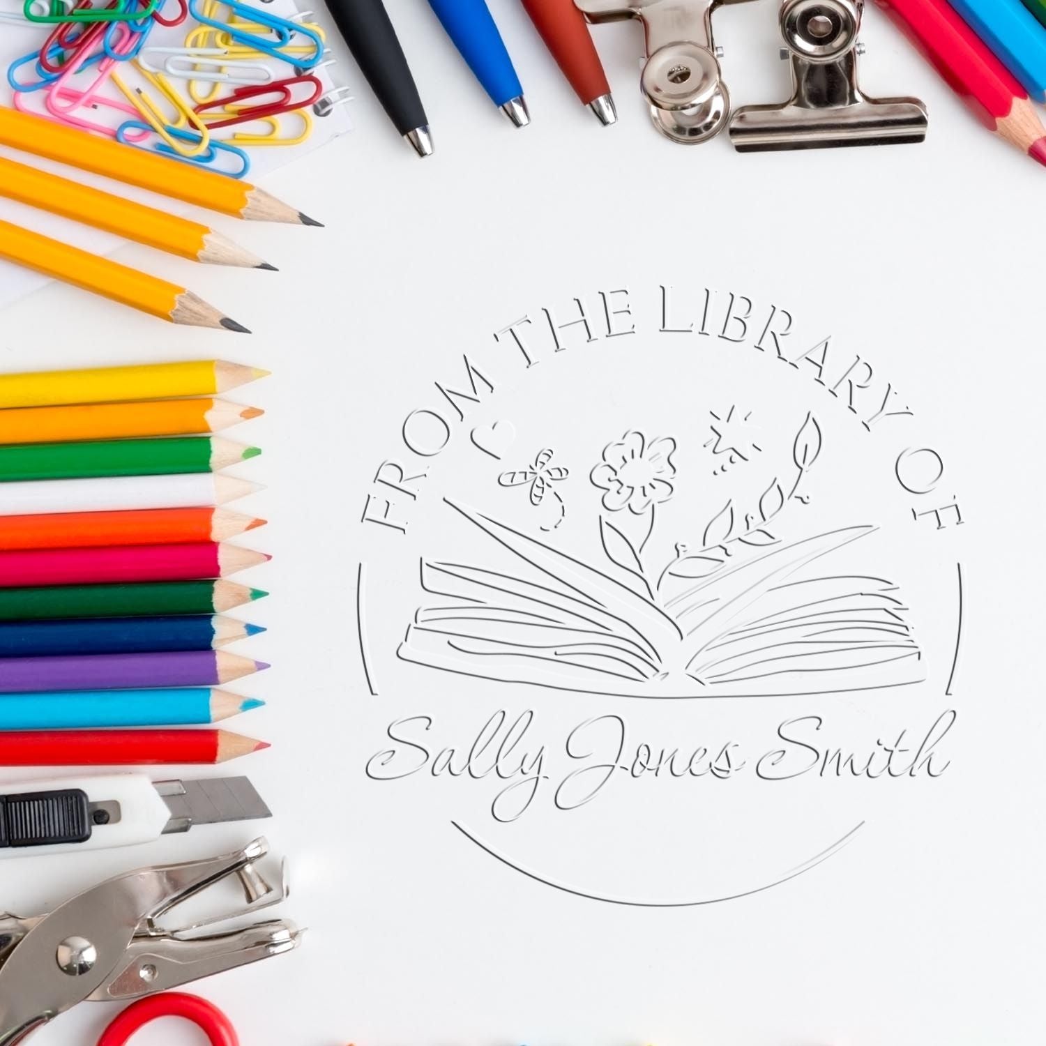 Circular silver library stamp seal reading From the Library of Sally Jones Smith, surrounded by colorful pencils, pens, and clips.