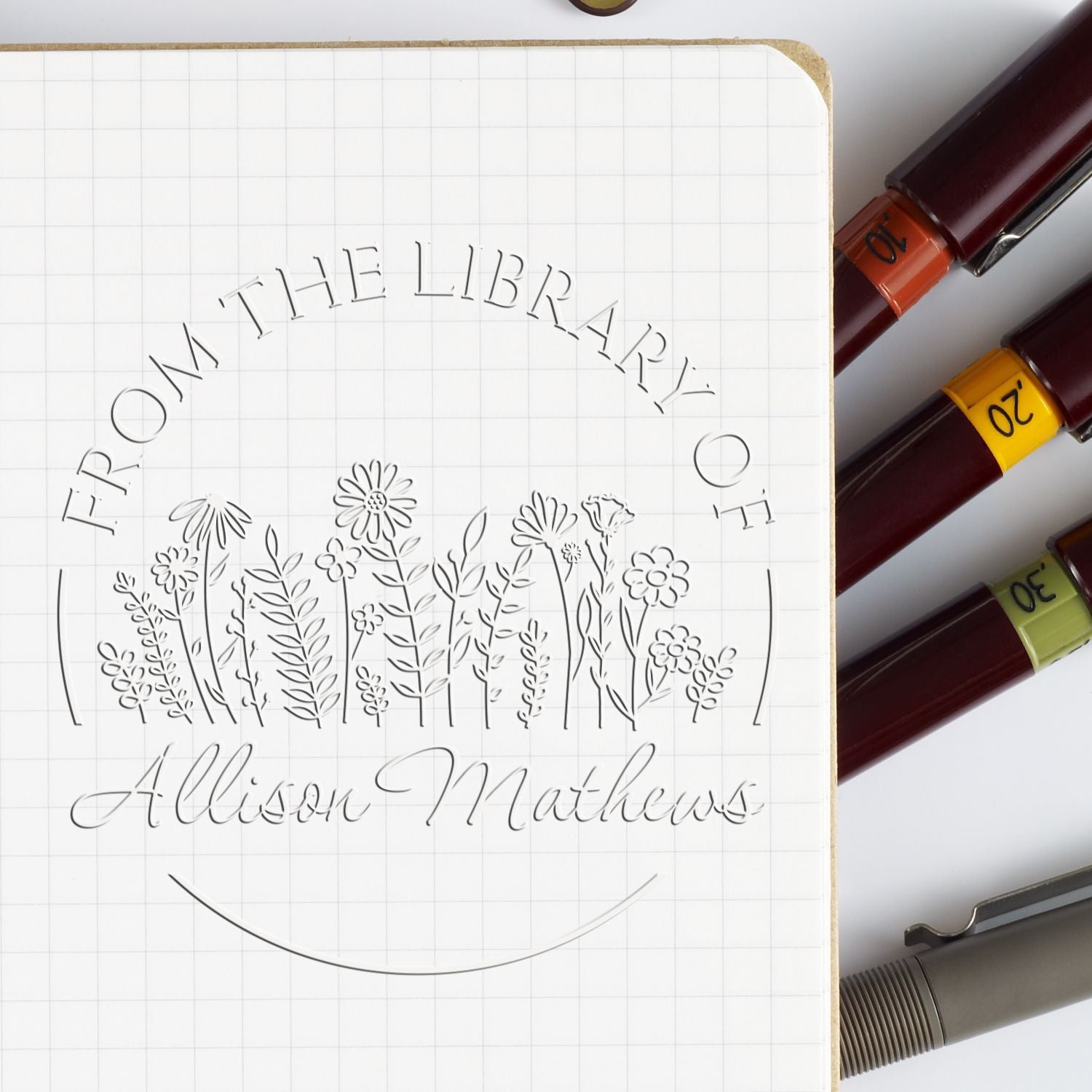 Library stamp seal on grid paper with floral line art, beside burgundy stamp markers with orange, yellow, and green bands.