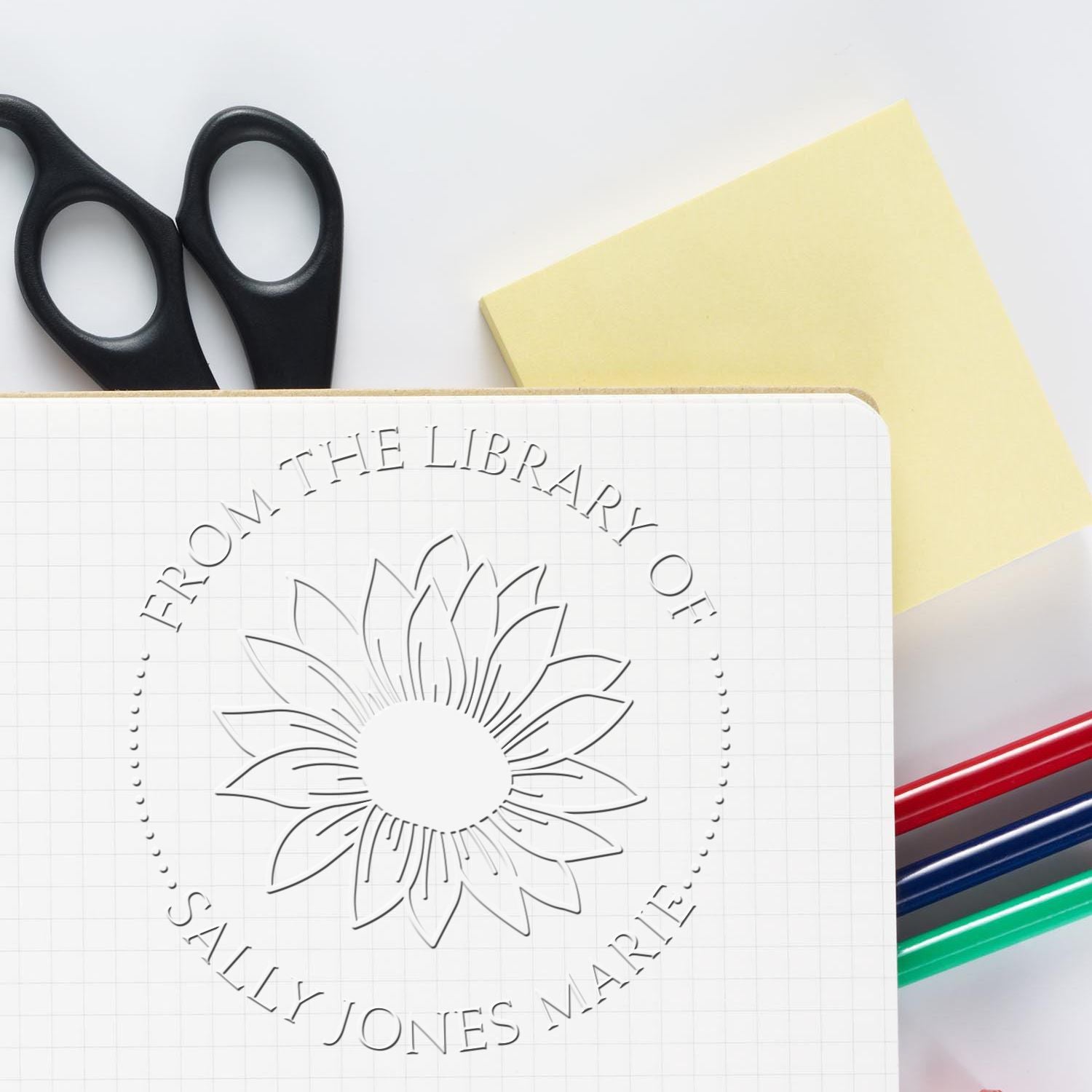 Custom embossed library seal on white grid notebook, with black scissors, yellow note, and red, blue, green pens