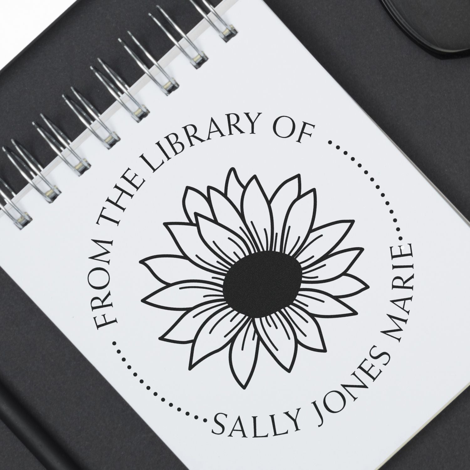 Black-and-white custom stamp and seal for The Library of Sally Jones Marie with sunflower logo on a spiral notebook.