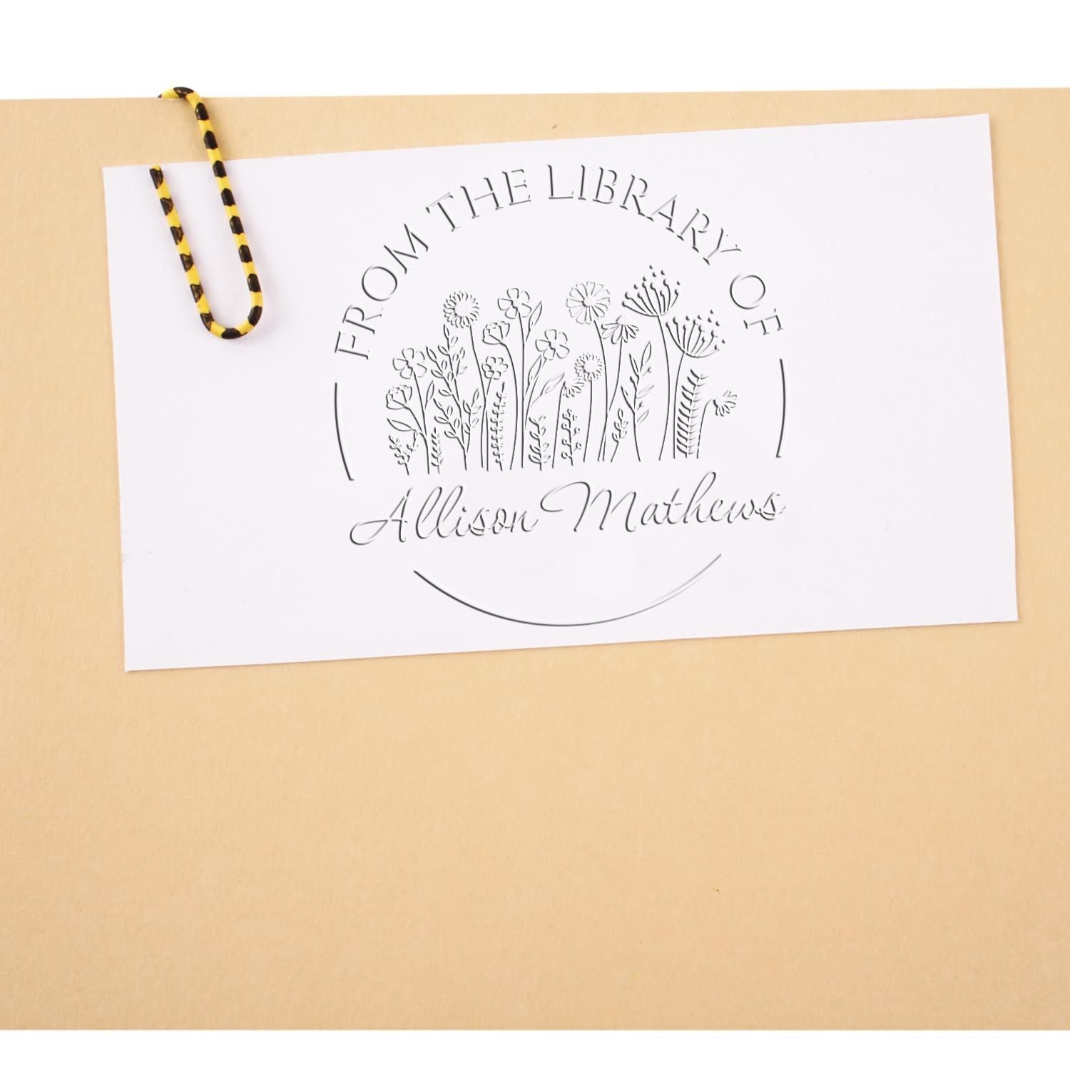 White custom stamp seal with floral library design and Allison Mathews text, clipped to a beige envelope with black-yellow cord.