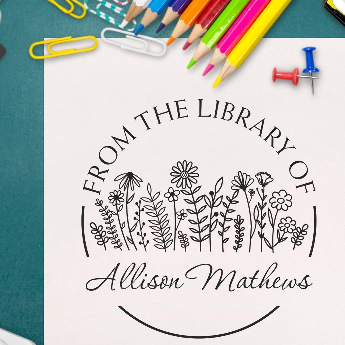 Custom library stamp and seal on white paper, black floral design and Allison Mathews text, with colorful pencils and pushpins.