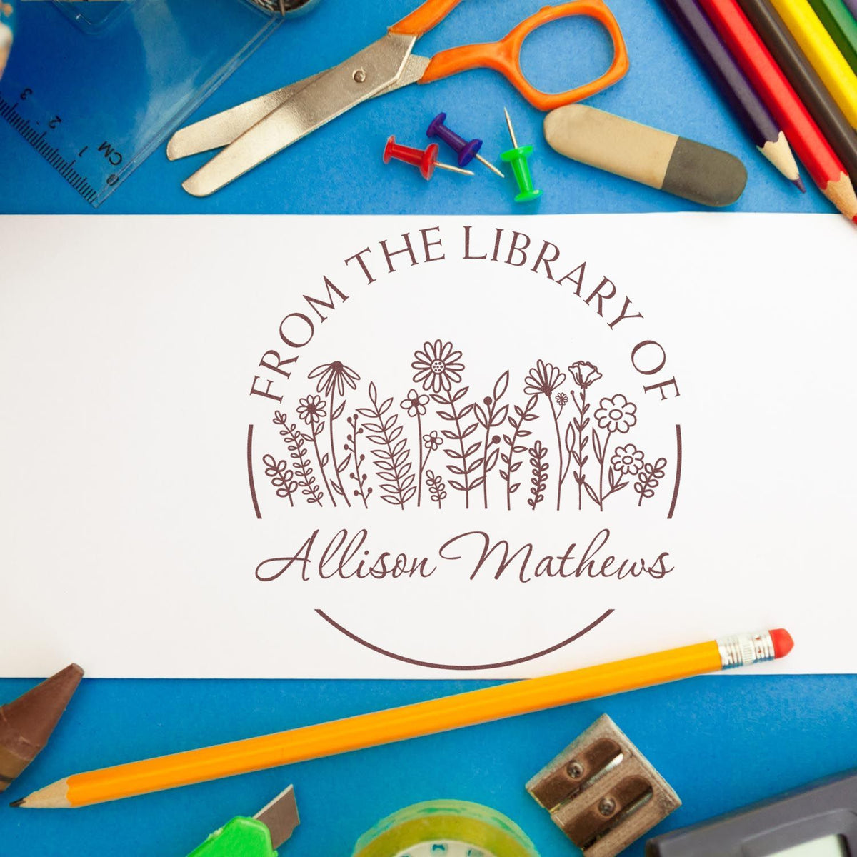 Personalized brown library stamp seal with floral design and Allison Mathews, surrounded by colorful school supplies on blue background.