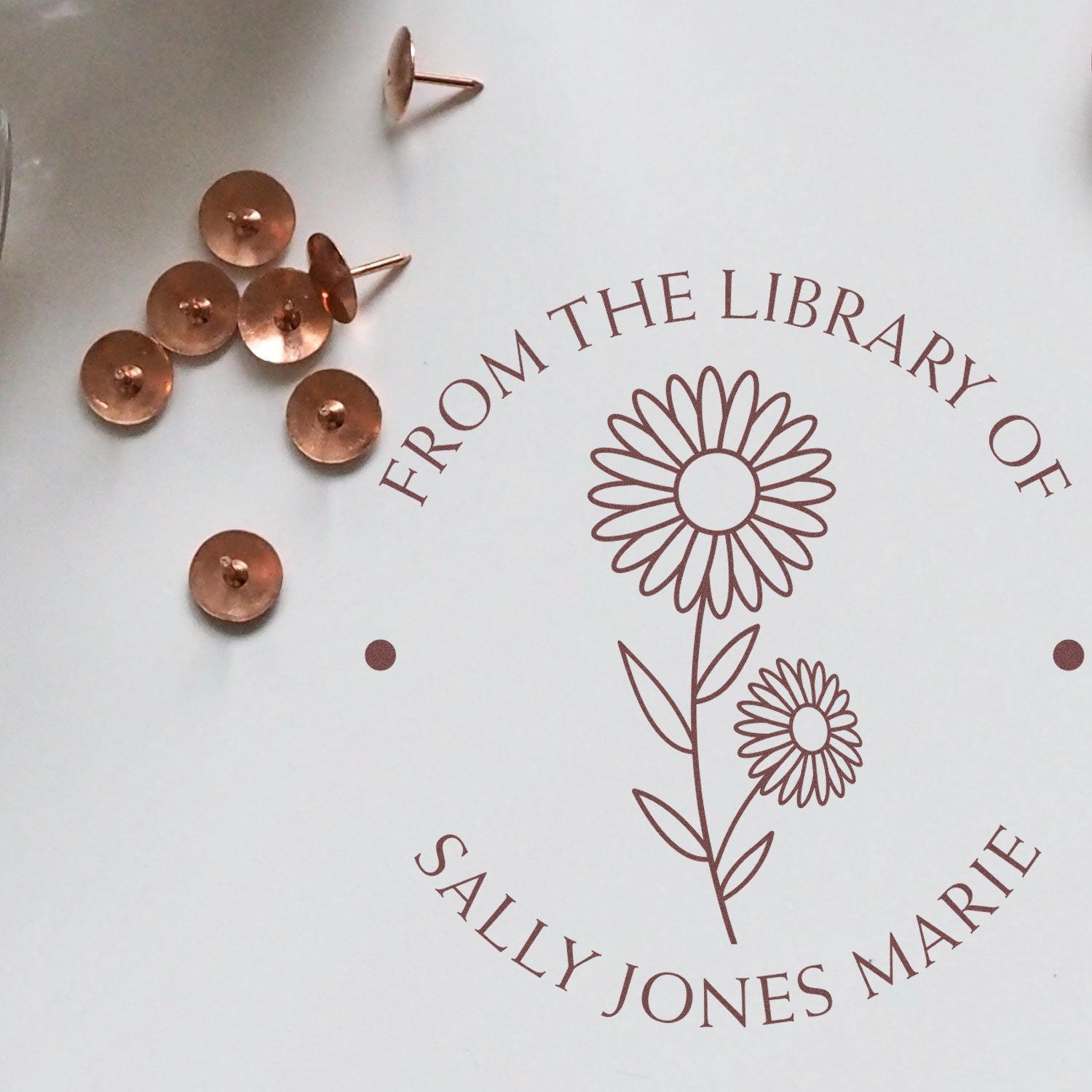 Copper stamp seals and fasteners beside a brown floral custom seal reading From the Library of Sally Jones Marie on white background.