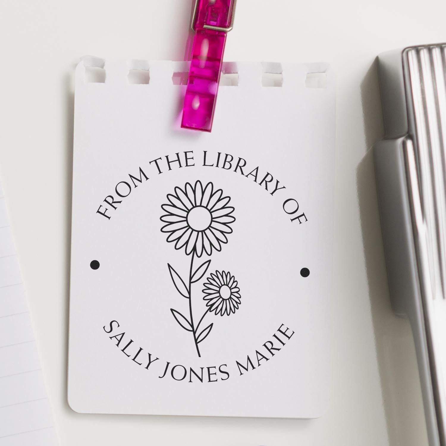 Personalized library stamp and seal card with black flower design, pink clip, and silver stamp block on white background.