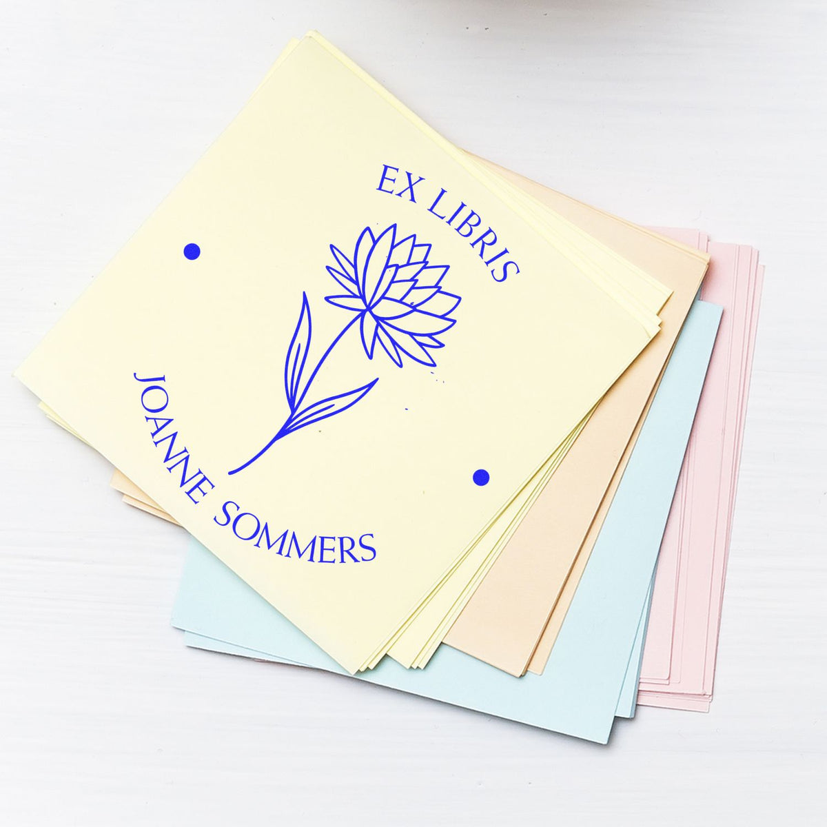 Pastel cardstock sheets with blue EX LIBRIS JOANNE SOMMERS floral book stamp seal, stacked on a white background.