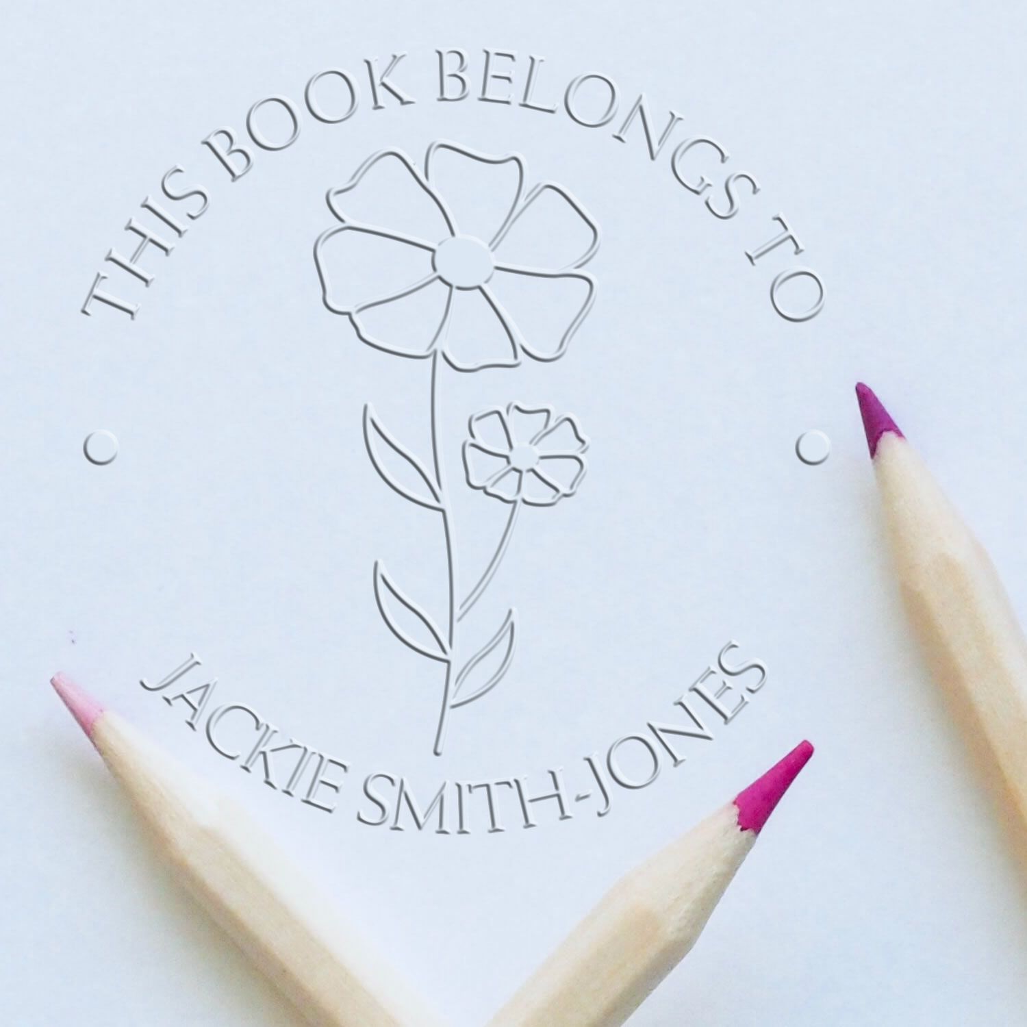 Custom embossed book stamp seal with floral design and pink pencils, reading This Book Belongs to Jackie Smith-Jones.