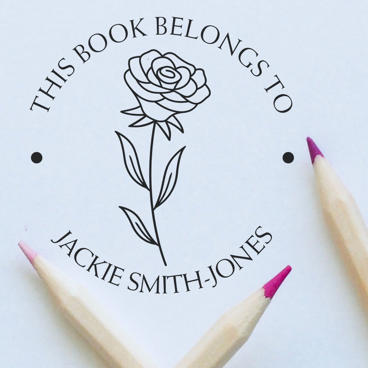 Custom black book stamp seal with rose outline and This Book Belongs to Jackie Smith-Jones on white paper, accented by pink pencil tips.