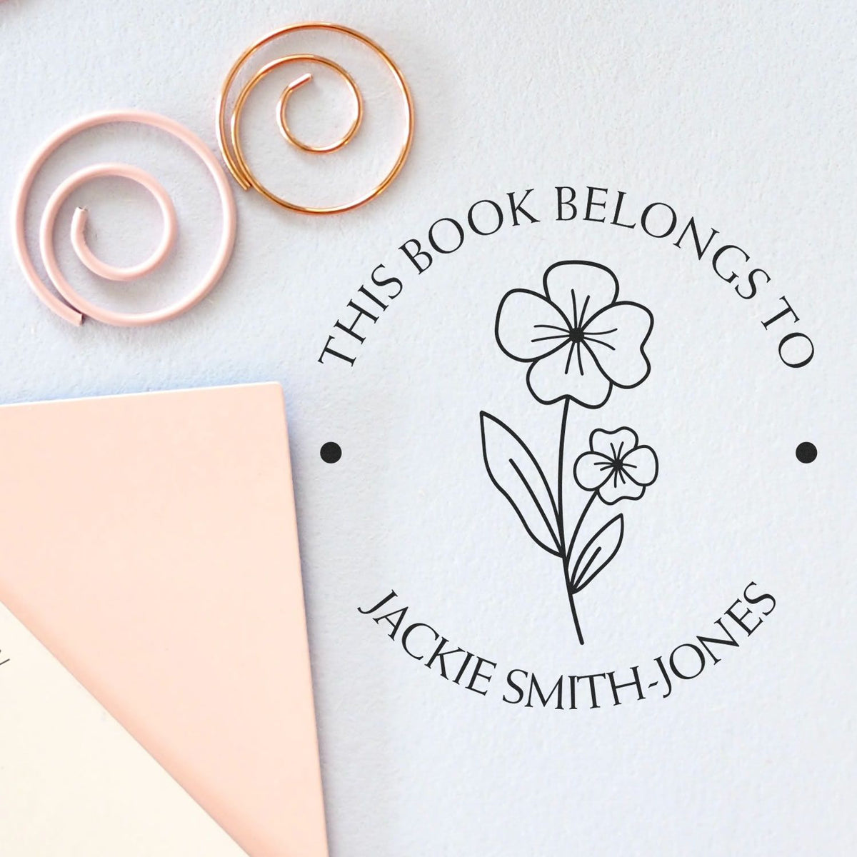 Custom floral book-belongs-to stamp in black on white paper, with pastel pink and gold wax seal swirls and pink envelope.