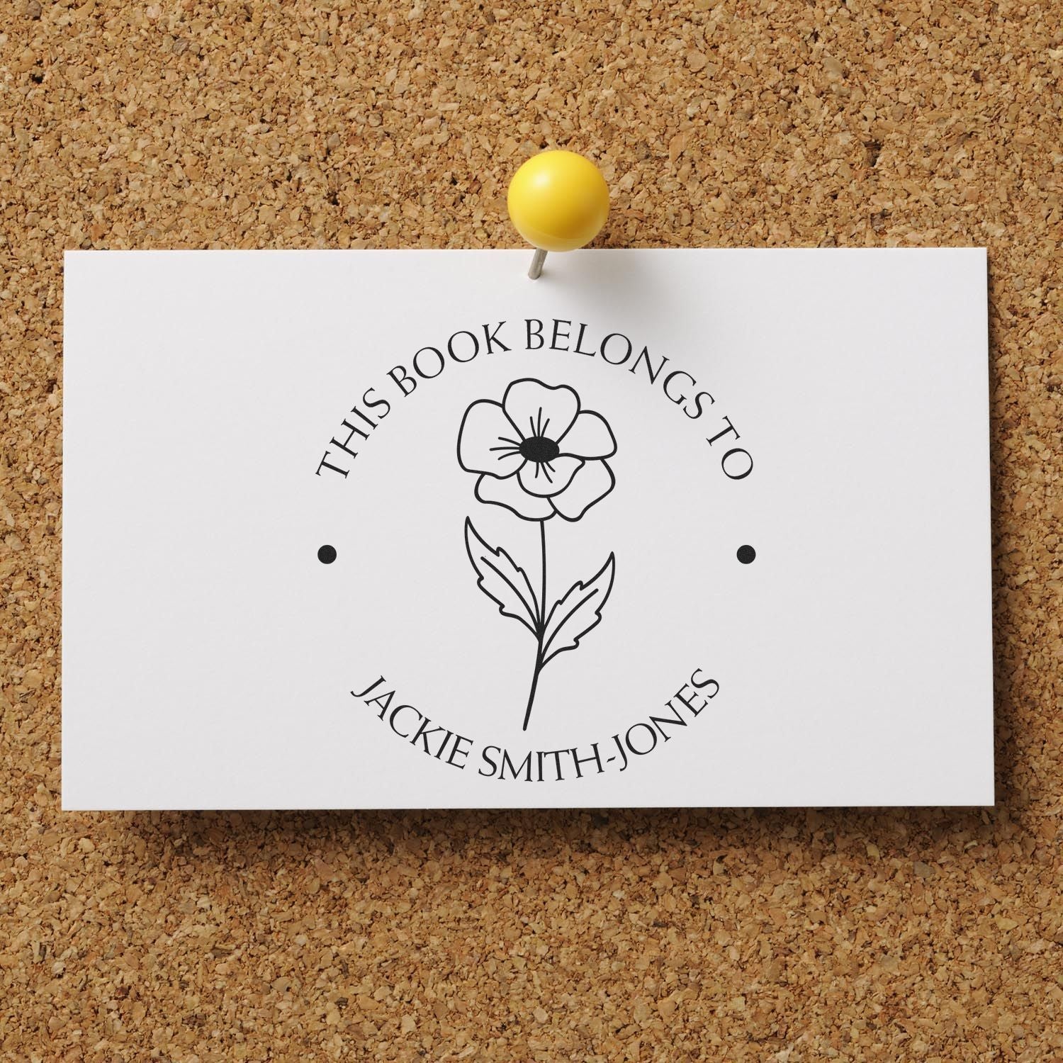 White personalized bookplate stamp with black floral seal, yellow pin, and corkboard background.