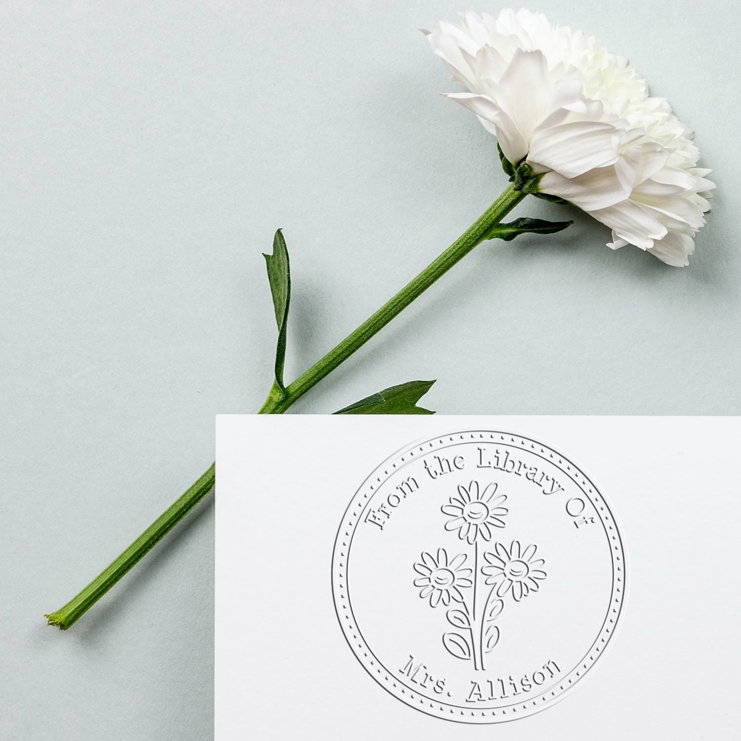 White flower beside a custom circular library seal stamp on white card, with a green stem on pale blue background.