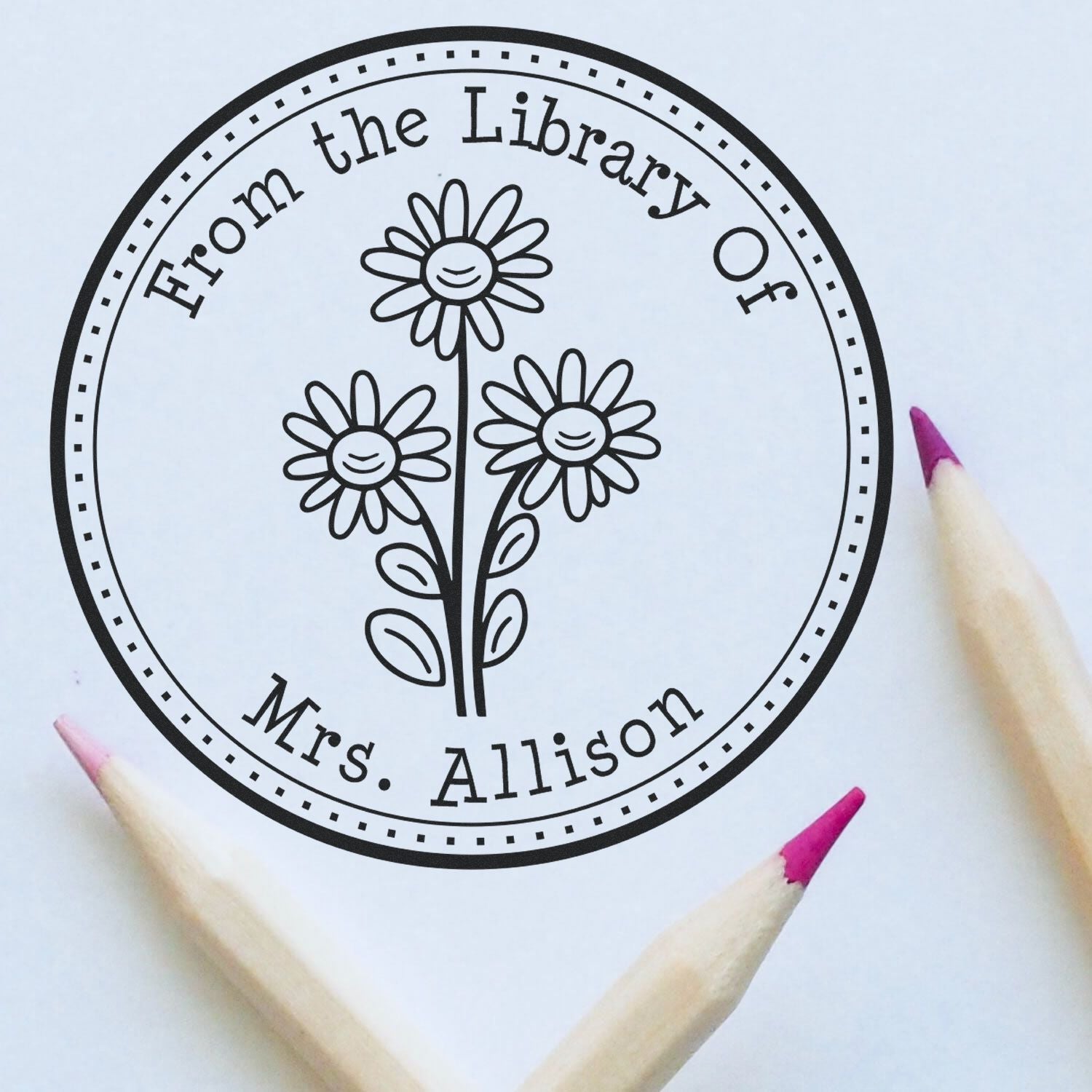 Black circular library stamp seal with smiling daisies and From the Library of Mrs. Allison text on white paper, pink pencils.