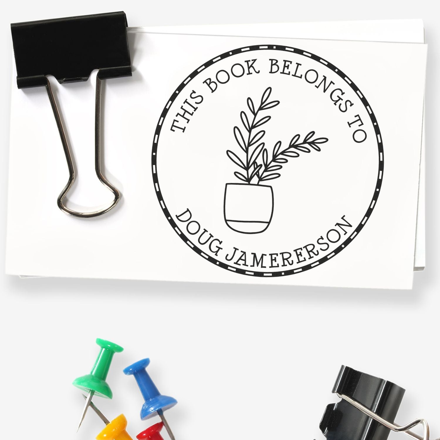 Black binder clip holding a white paper book stamp and seal with circular This Book Belongs to Doug Jamerson plant design.