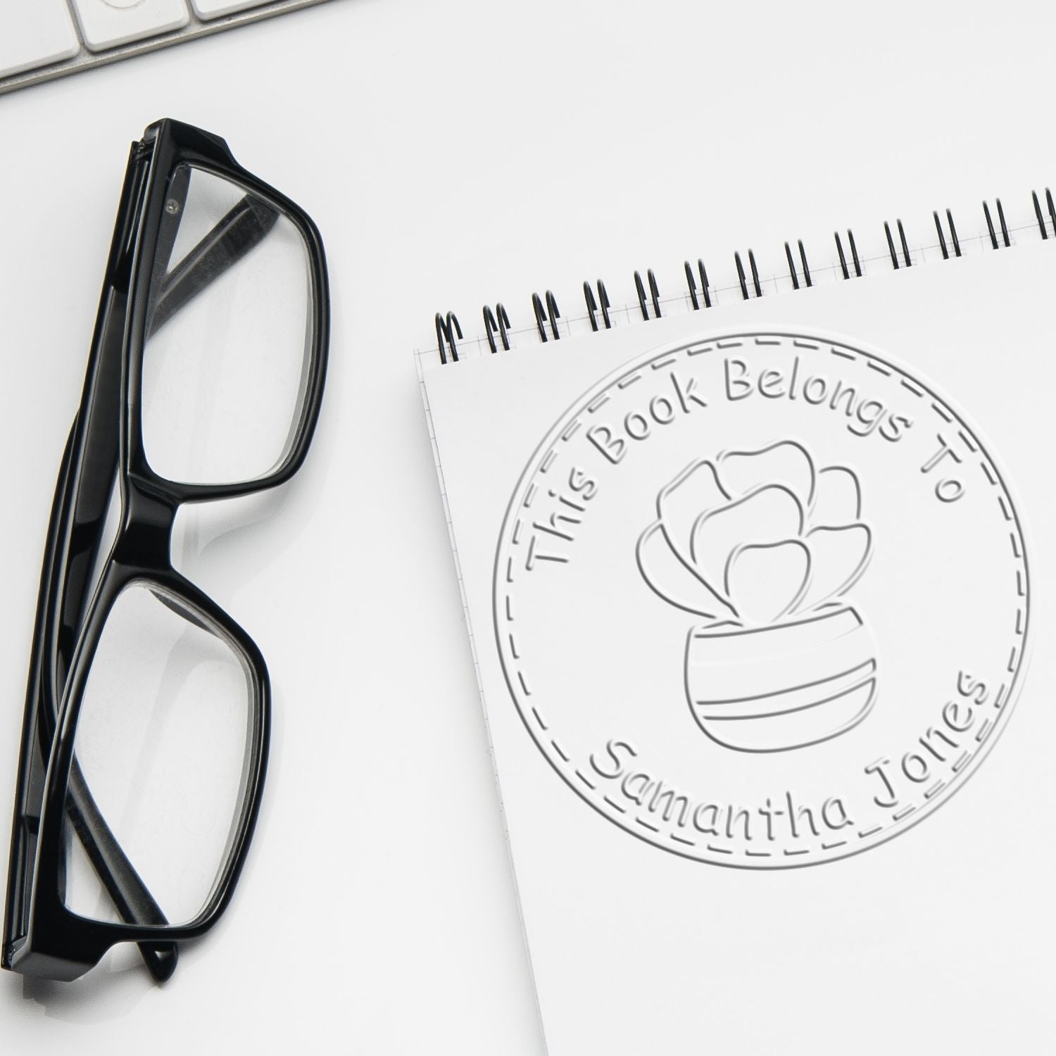 Black eyeglasses beside a spiral notebook with a custom embossed library book ownership seal and flower pot design.