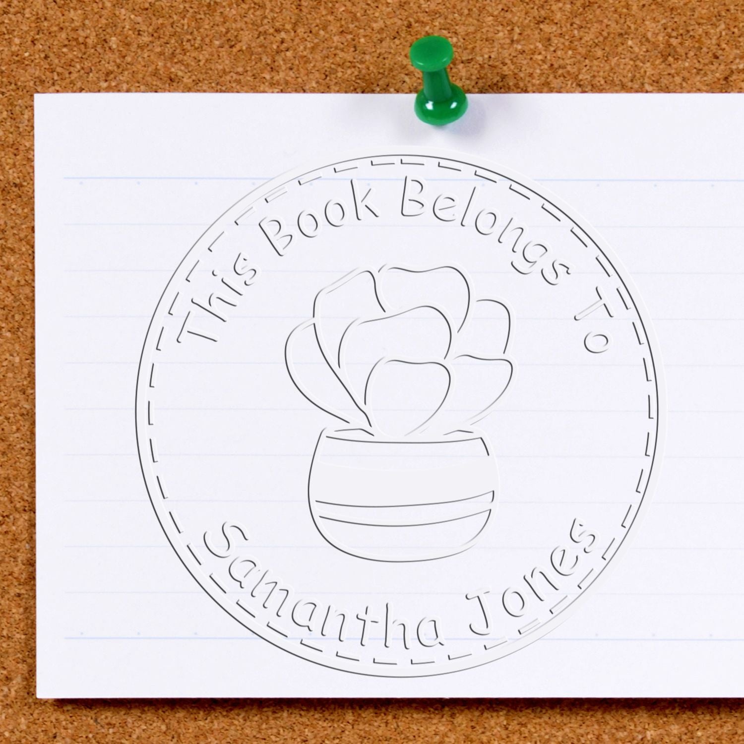 White bookplate stamp seal on lined paper, pinned with a green tack, embossed with a potted plant and This Book Belongs to Samantha Jones.