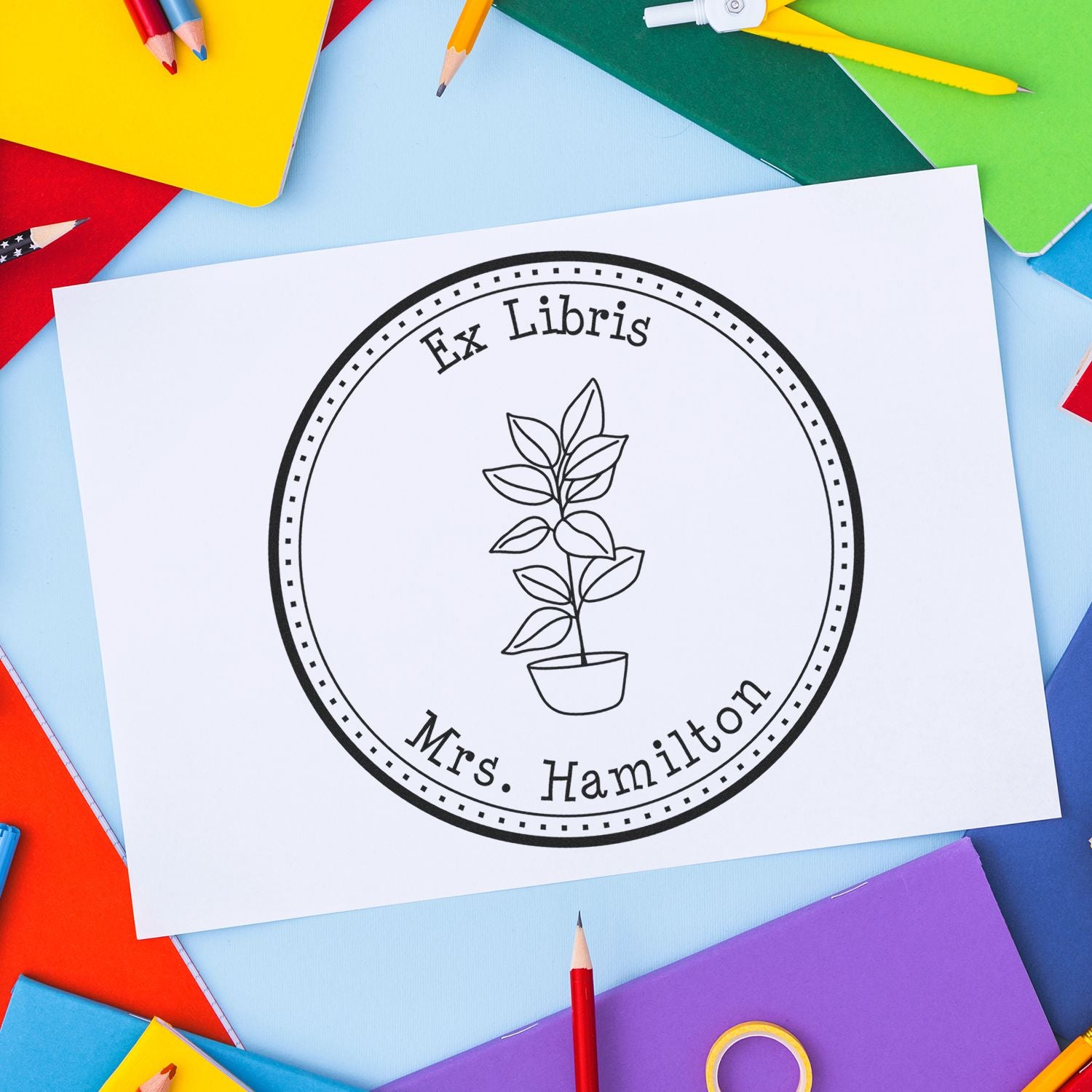 Custom black Ex Libris stamp seal with potted plant on white paper, surrounded by colorful pencils and notebooks.