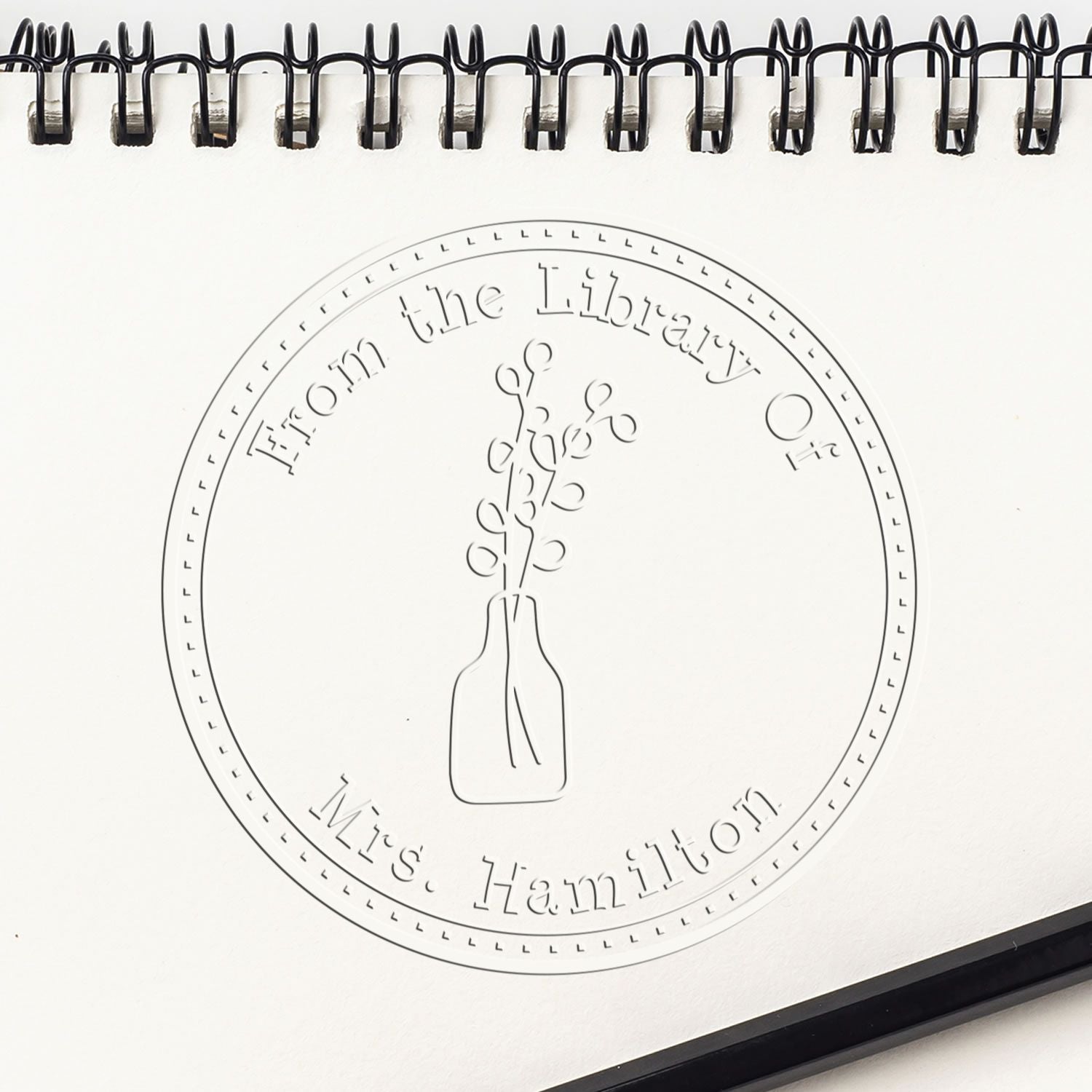 White spiral notebook featuring a black embossed library seal stamp with circular border and vase of flowers, Mrs. Hamilton.