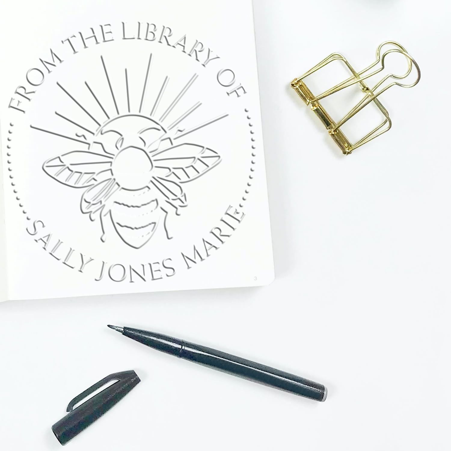 Custom library stamp and seal proof with silver bee design, gold binder clip, and black pen on a white background.