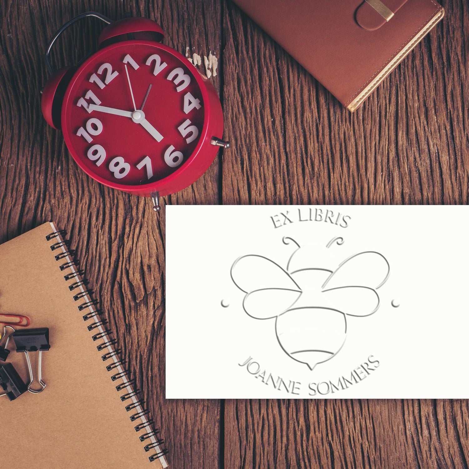 Custom stamp and seal with embossed bee logo on white card, beside a red alarm clock and tan notebooks on wood