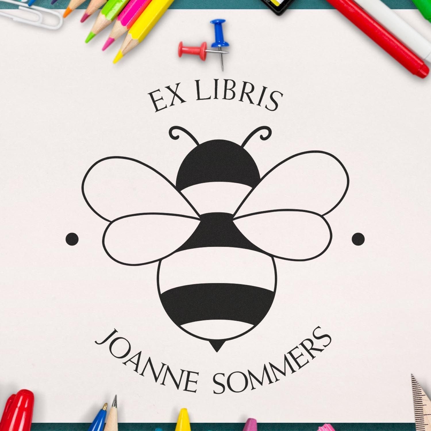 Black bee ex libris book stamp seal with Joanne Sommers, framed by bright colored pencils and push pins on white.