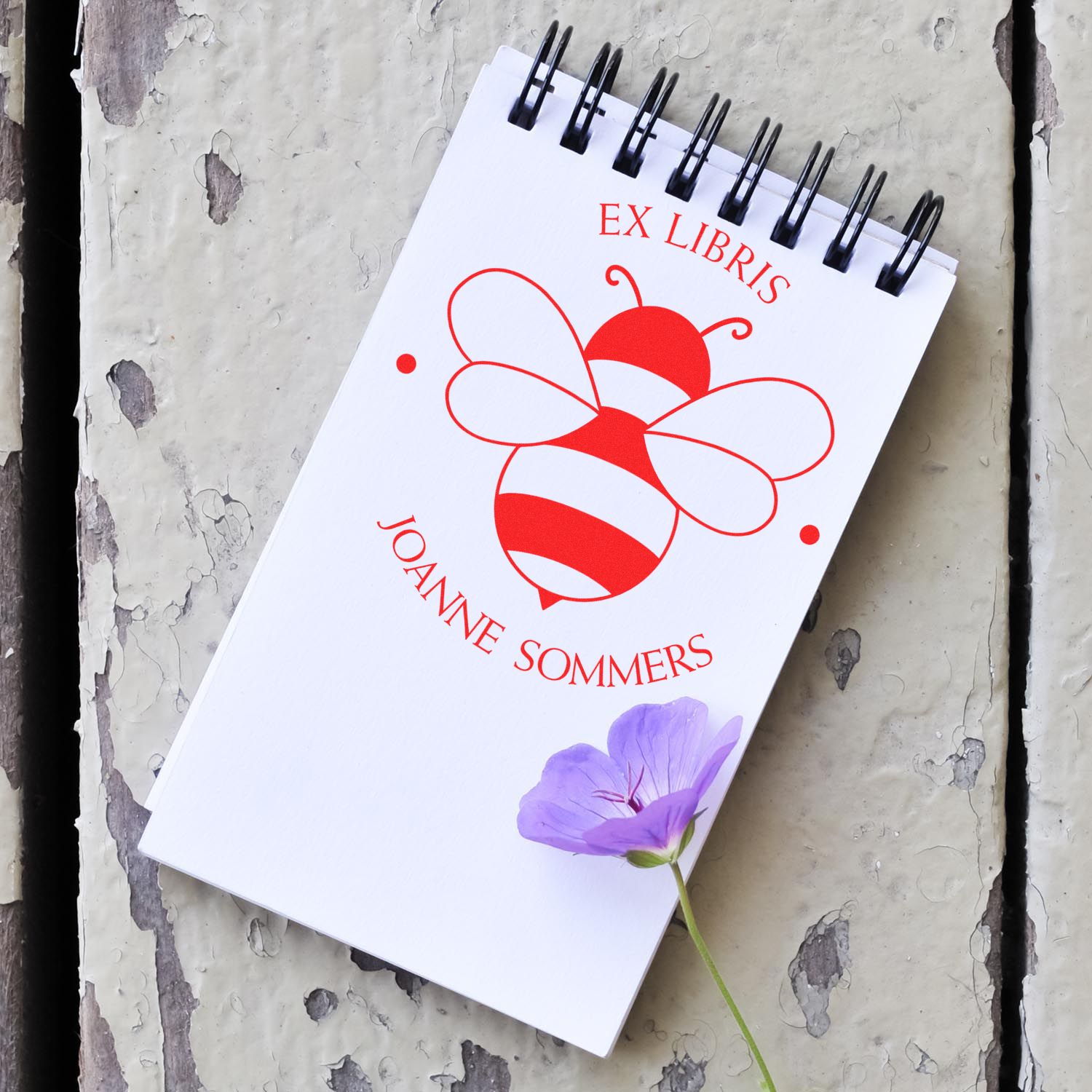 White spiral notebook with red ex libris bee stamp and seal design, purple flower, on weathered gray wood.