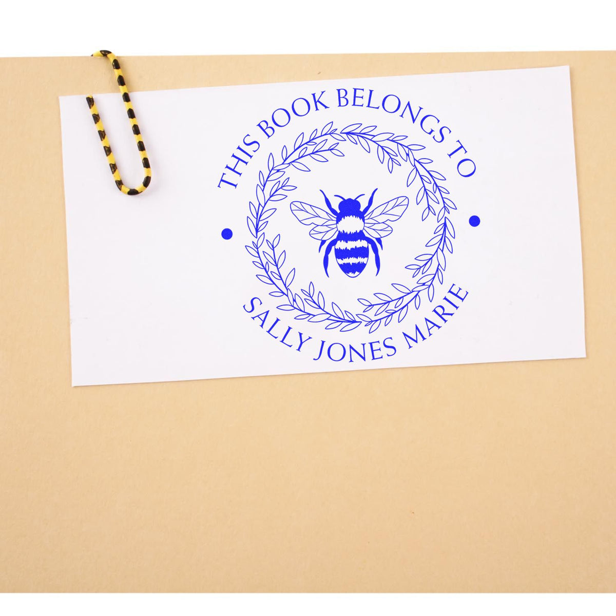 Blue custom book stamp and seal with bee and leafy wreath, reading This Book Belongs to Sally Jones Marie on white card.