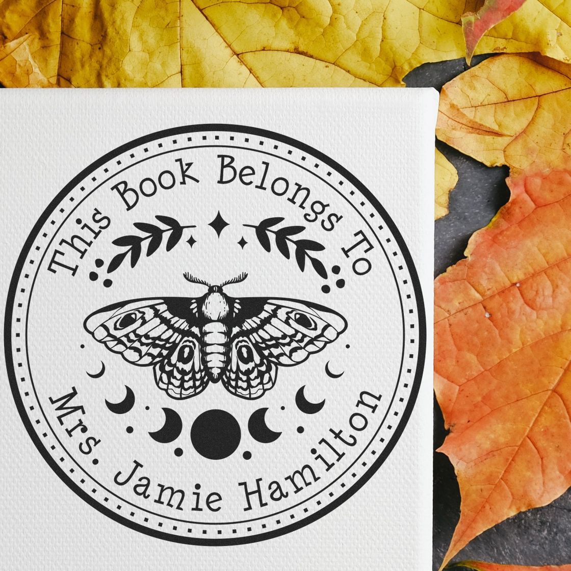 Black custom bookplate stamp and seal with moth, crescent moons, and Mrs. Jamie Hamilton on white paper, framed by autumn leaves.