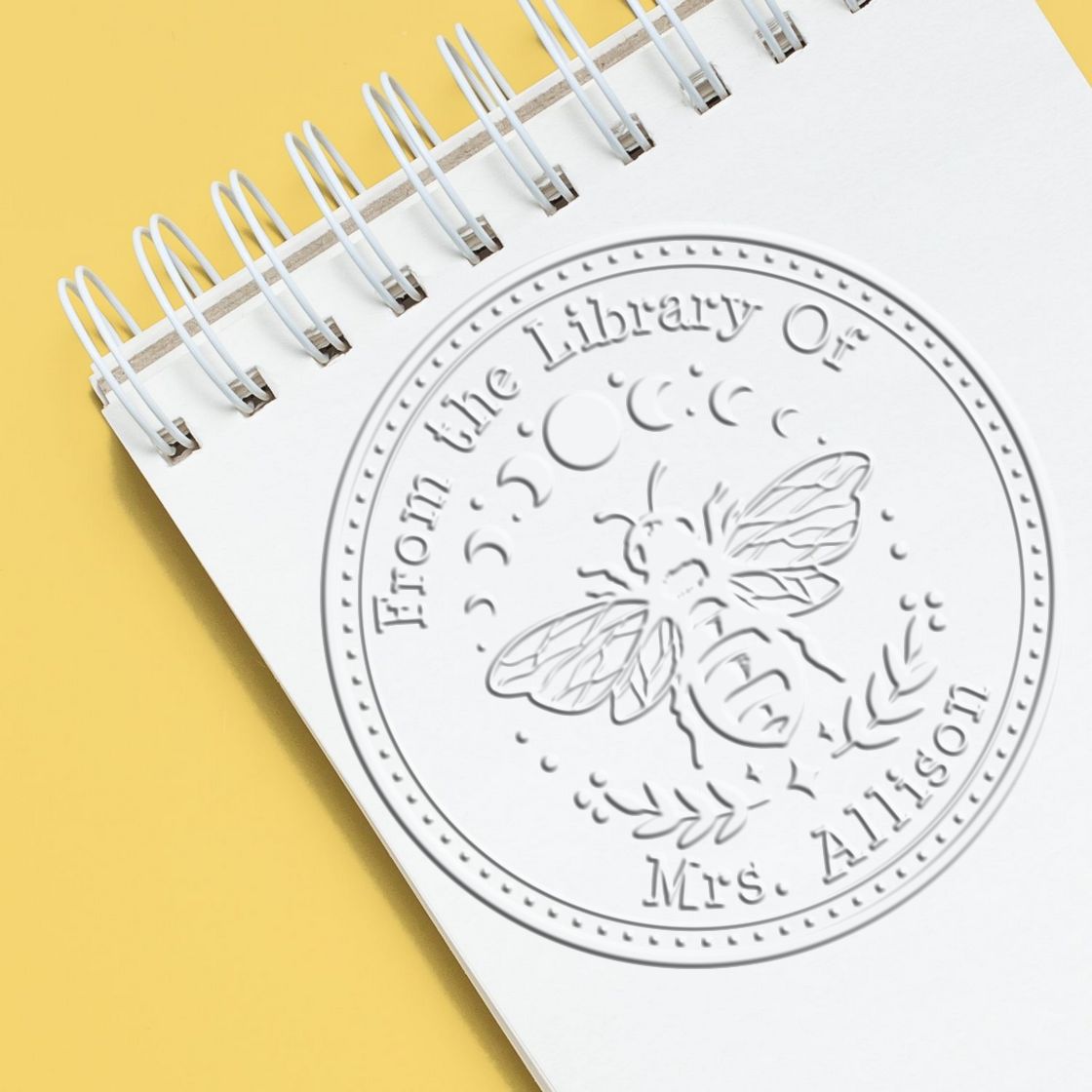 Embossed white circular library stamp seal with bee and From the Library Of on a spiral notebook over a yellow background.
