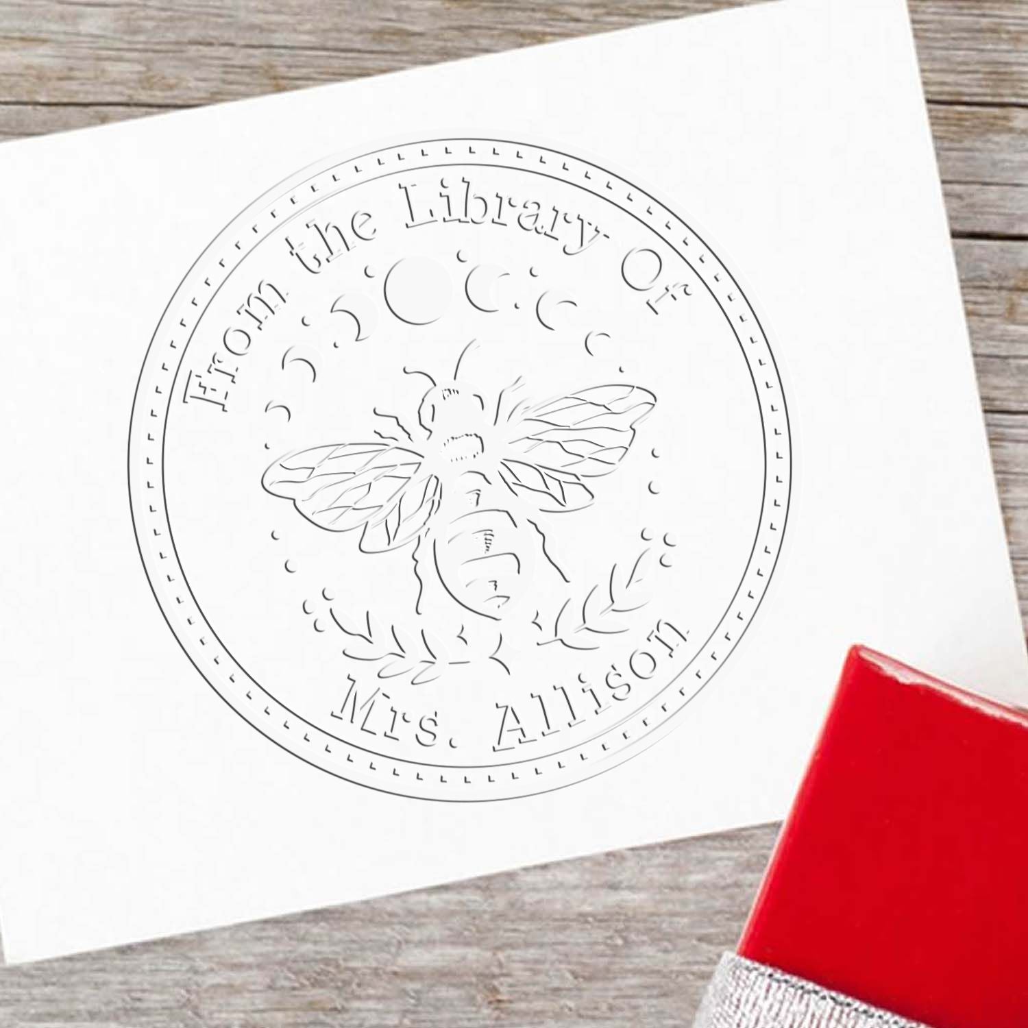 Custom bee-themed library embossing seal reading From the Library Of Mrs. Allison on white paper, with a red stamp handle.