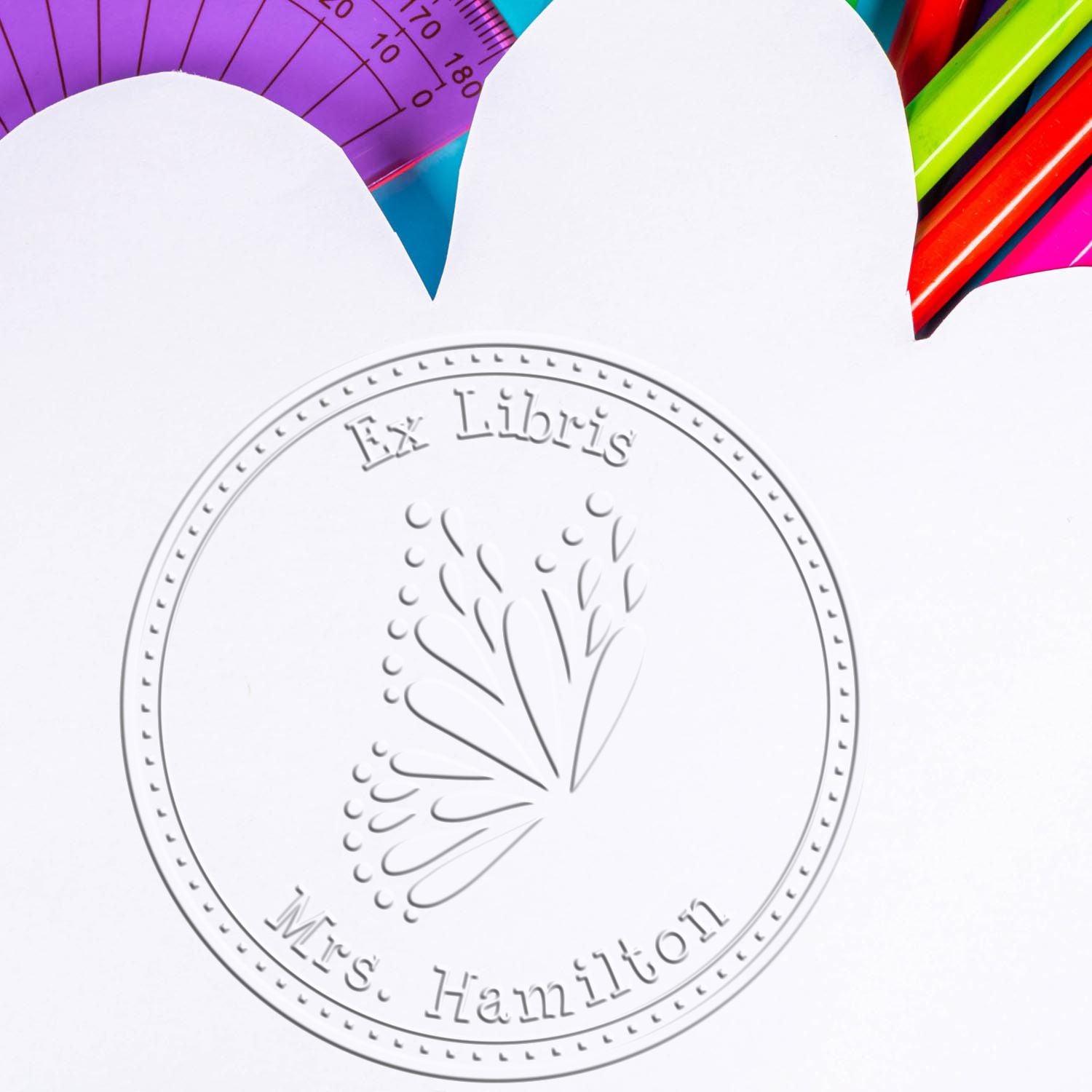 Embossed white stamp seal with Ex Libris Mrs. Hamilton beside colorful pencils and a purple ruler.