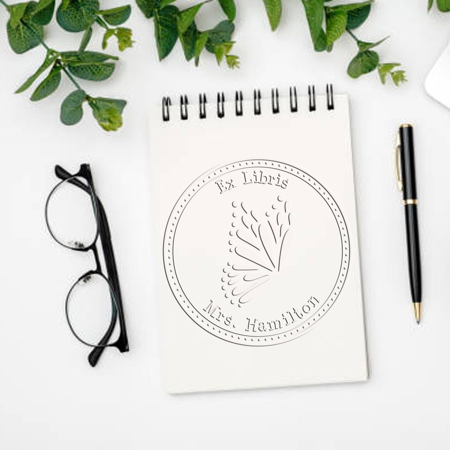 Personalized round stamp seal on white spiral notebook, with black glasses, pen, and green leaves on a clean desk.