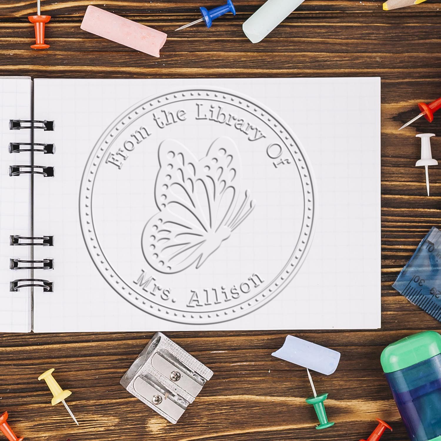 Round white embossed library stamp seal with butterfly on notebook, on brown wood desk with colorful push pins.