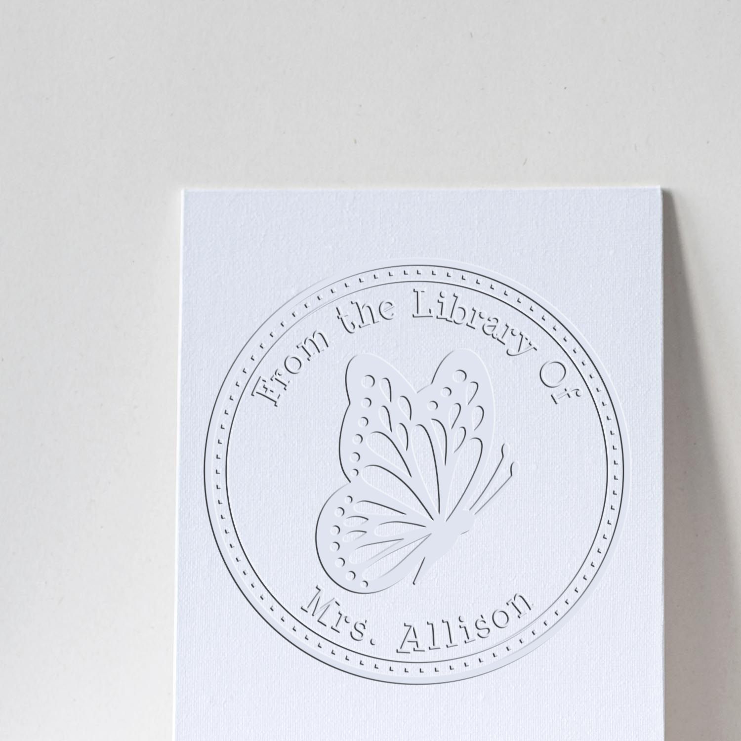 White embossed circular library stamp seal with butterfly on textured white paper, reading From the Library of Mrs. Allison.