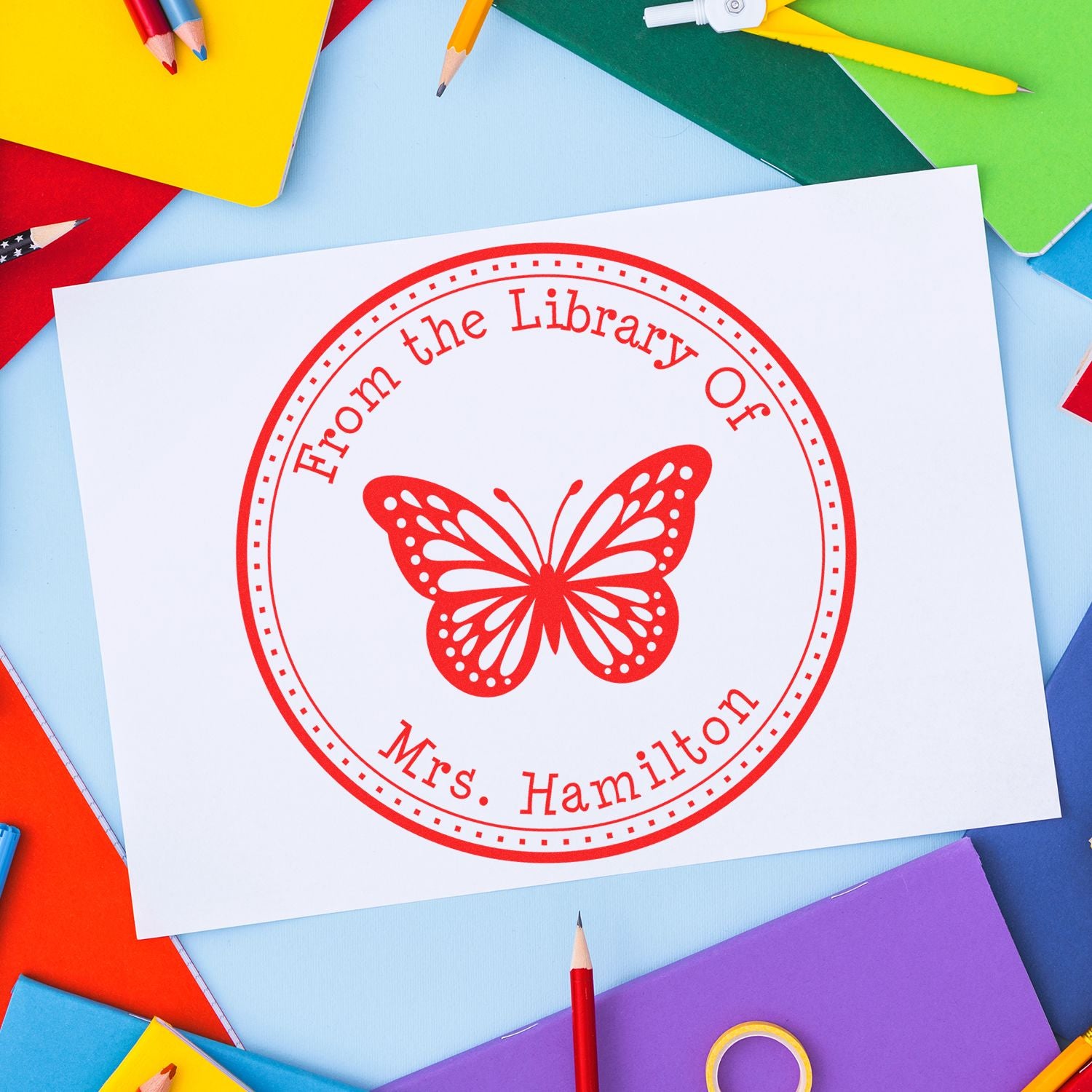 Red butterfly library stamp seal on white paper, with colorful notebooks and pencils around it.