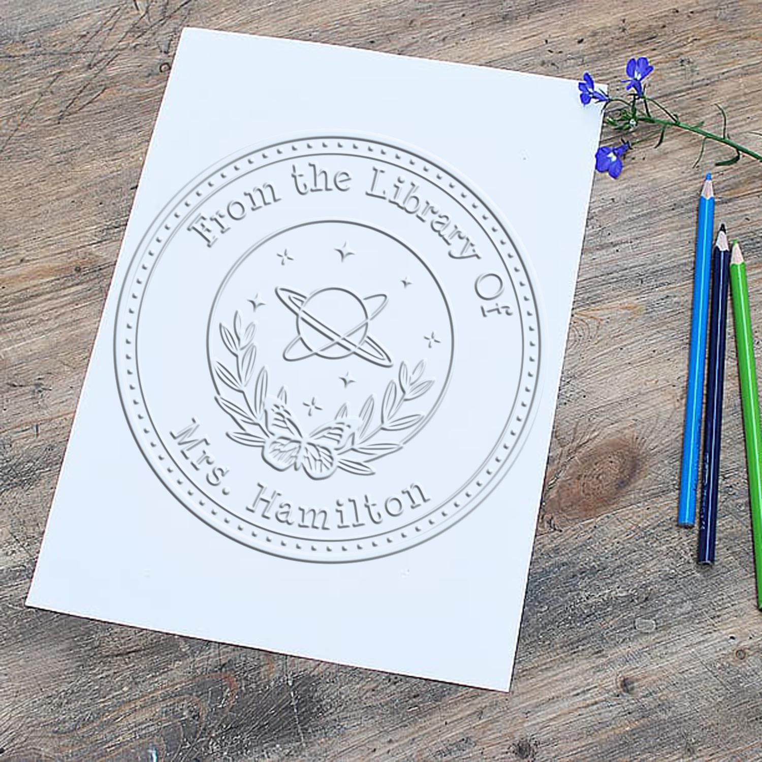 White paper with embossed circular library seal stamp reading From the Library of Mrs. Hamilton on a wood desk.