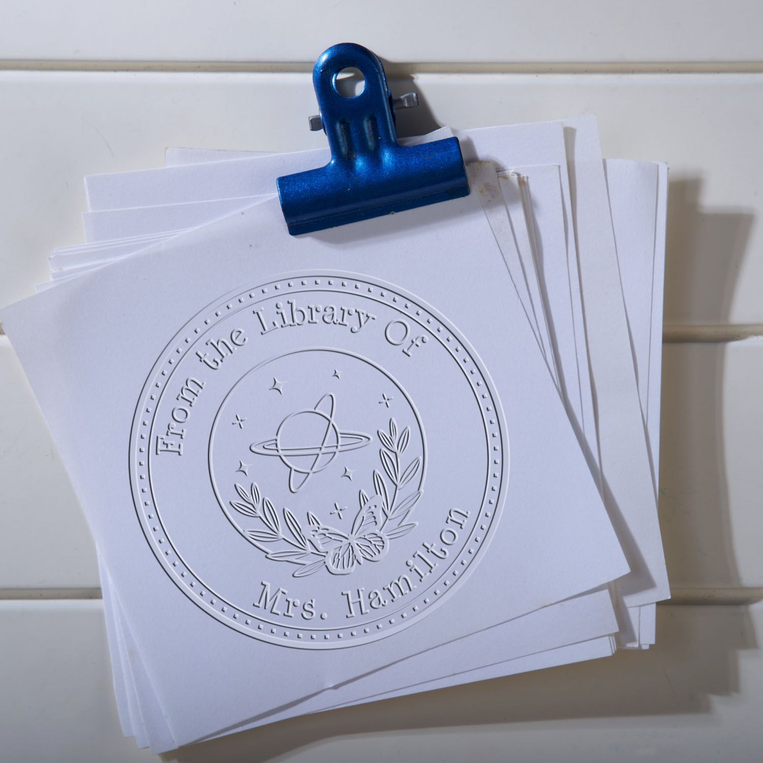 Blue binder clip holding white stamp and seal sample sheets with embossed circular library seal, wreath, and butterfly design.