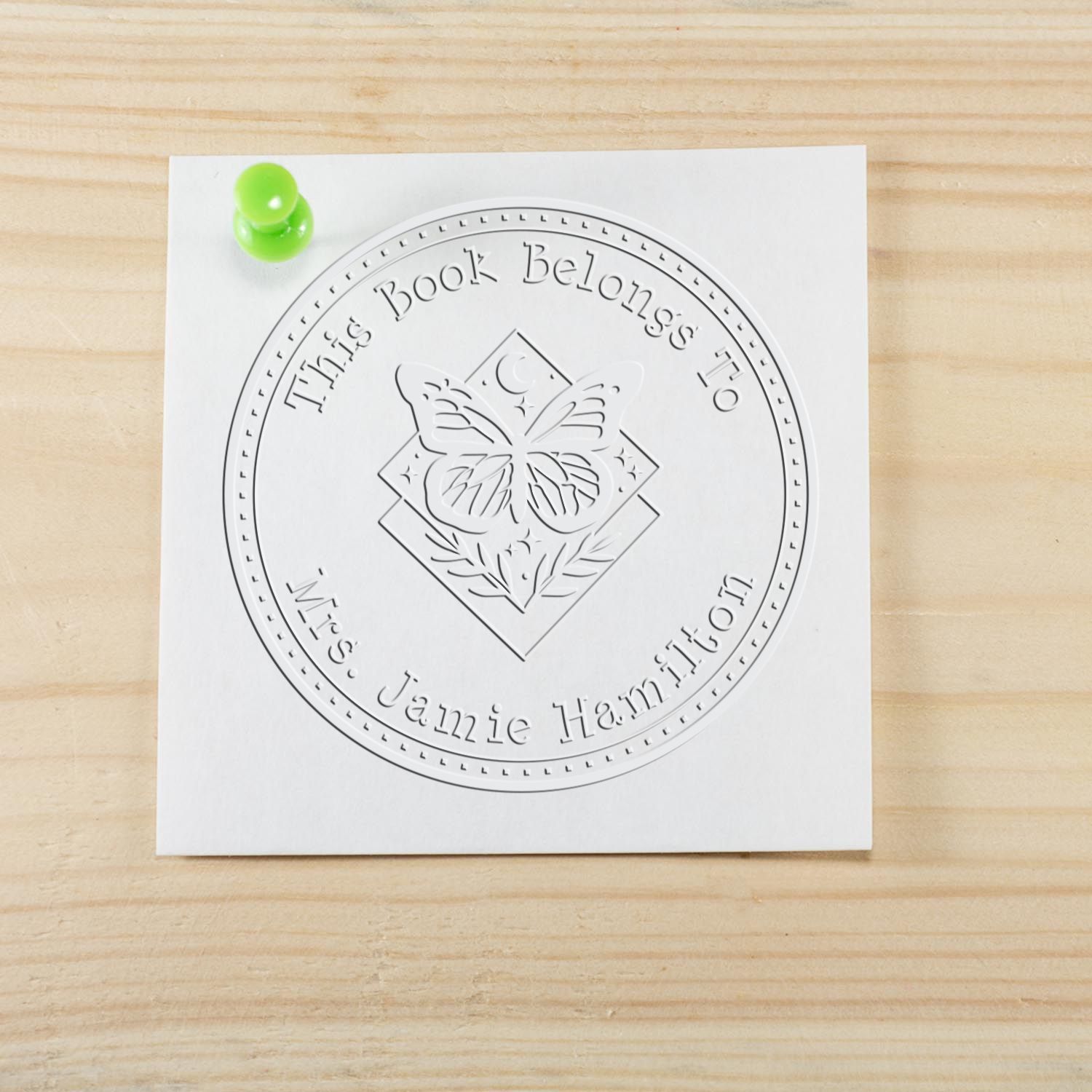 White custom book stamp seal with butterfly design, green pushpin, and embossed text This Book Belongs to Mrs. Jamie Hamilton.