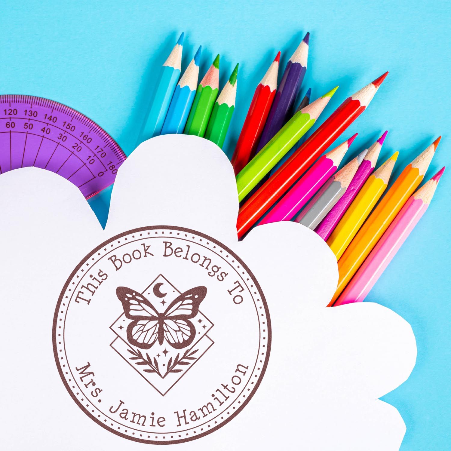 Personalized brown book ownership stamp and seal with butterfly on white cloud cutout, framed by colorful pencils and a purple protractor.