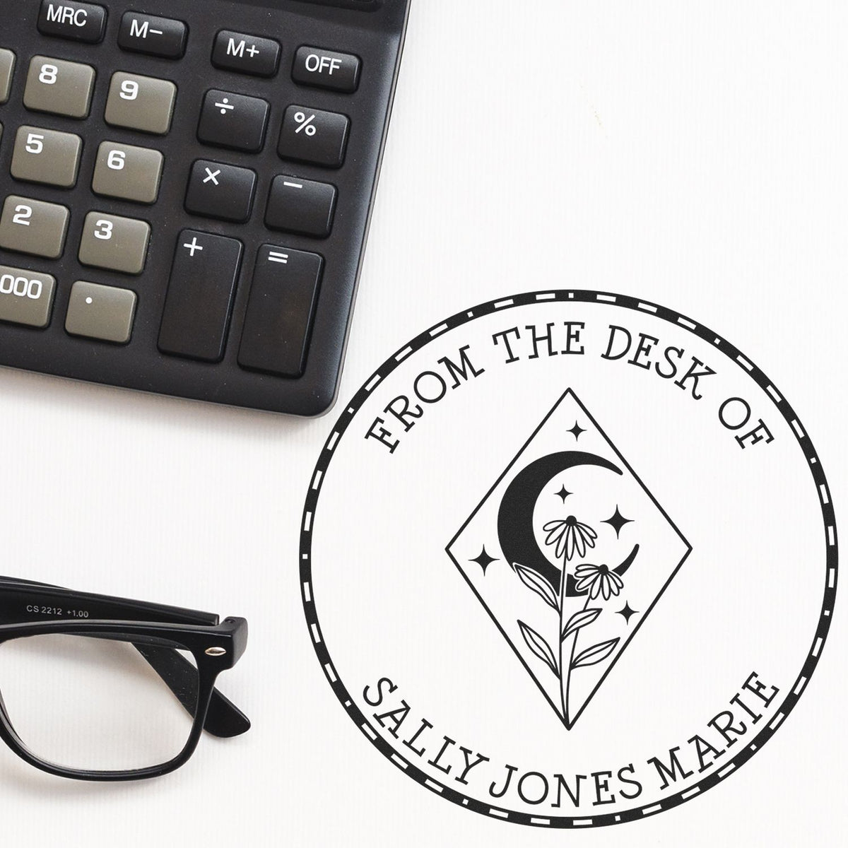 Black custom stamp seal with crescent moon, flowers, and white circular text beside a black calculator and glasses.
